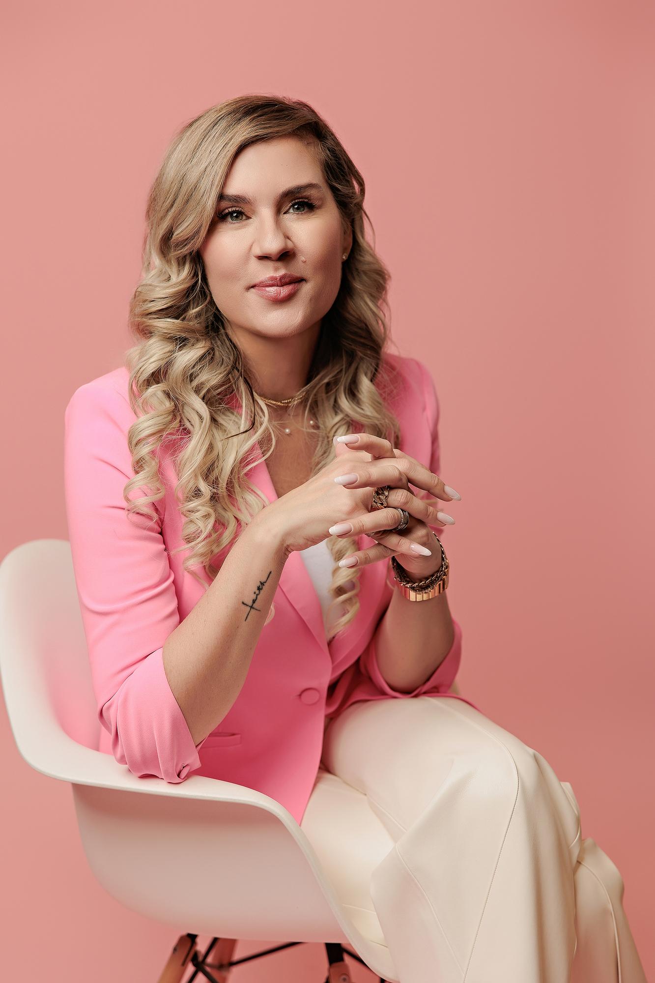 Corporate headshot photography Calgary: Polished woman in a hot pink blazer and cream trousers seated on a white chair with hands clasped, soft pink background