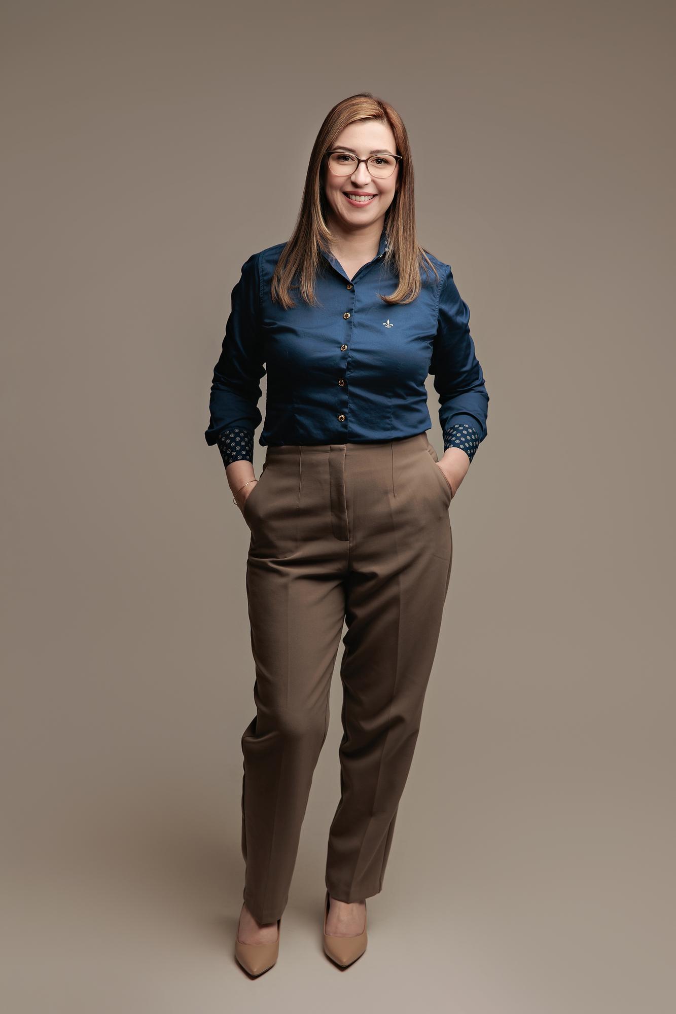 Full-length professional headshot Calgary: Smiling woman with glasses in a navy button-up shirt and taupe high-waisted trousers standing with hands in pockets, warm taupe background