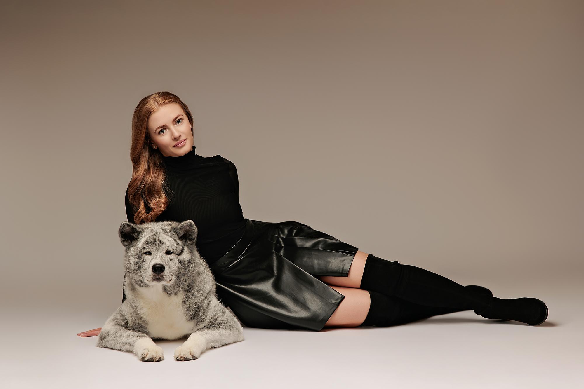 Personal branding photography Calgary studio: Editorial portrait of a woman with long auburn wavy hair in a black turtleneck and black leather skirt, reclining elegantly on the studio floor beside a large grey-and-white Akita dog against a warm taupe background