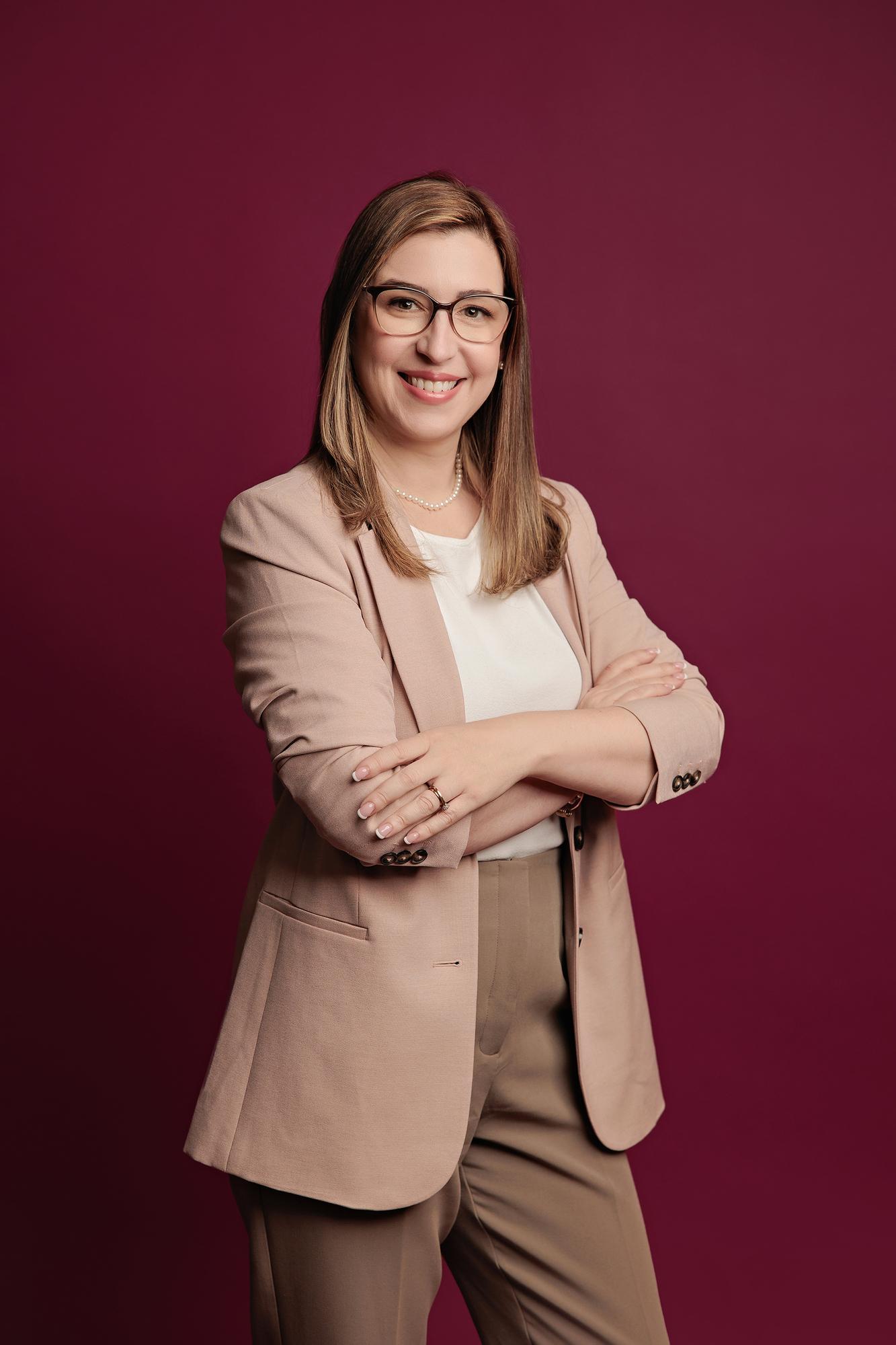 Corporate headshot photography Calgary: Confident woman with glasses in a blush blazer and white top standing with arms crossed and a warm smile against a deep burgundy background