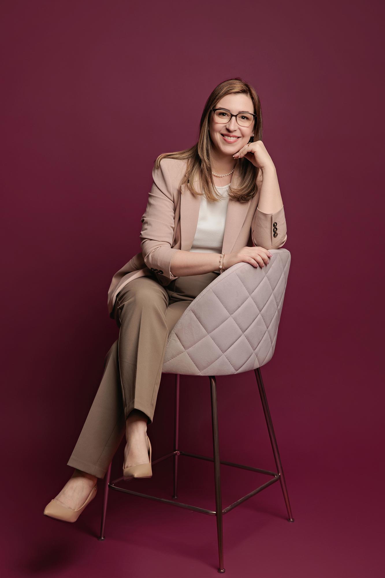 Professional headshot Calgary studio: Approachable woman with glasses in a blush blazer and taupe trousers seated on a quilted velvet chair resting her chin on her hand, burgundy background