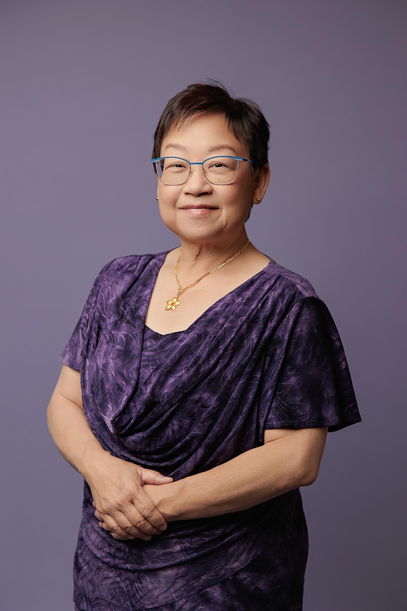 Professional headshot Calgary: Gracious senior professional woman with blue-frame glasses in a purple jacquard cowl-neck top and gold flower necklace, hands clasped, purple background