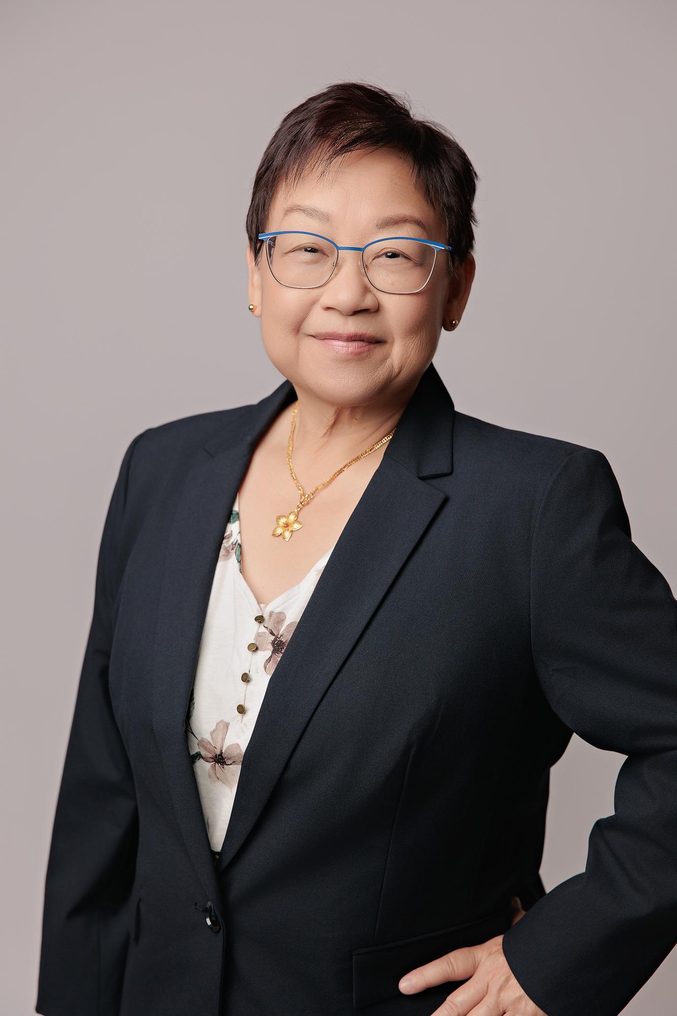 Executive headshot photography Calgary: Distinguished senior professional woman with blue-frame glasses in a navy blazer and floral blouse standing with hand on hip, light grey background