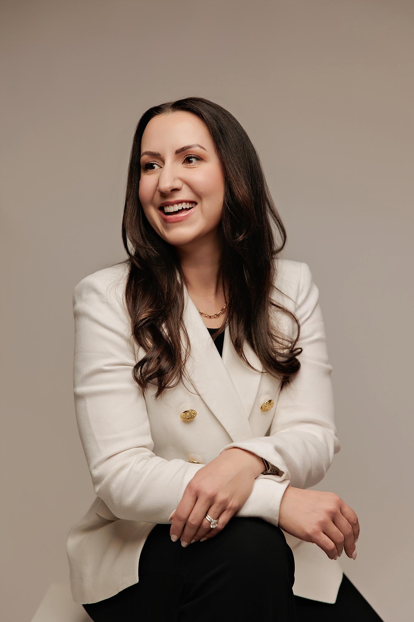 A woman with long dark hair smiles in a white blazer during a Calgary professional headshot session, captured with soft studio lighting and a neutral backdrop.