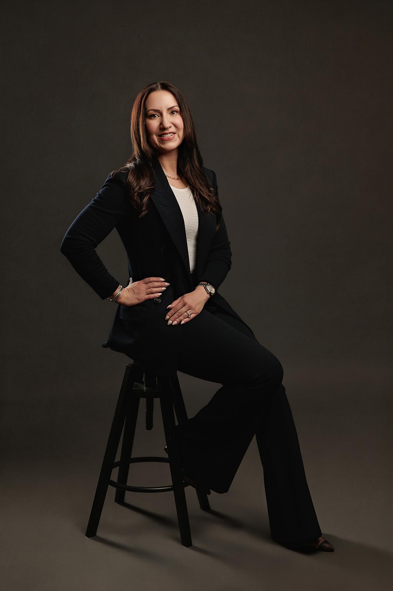 A professional woman in a navy suit poses confidently on a stool in our Calgary photography studio, showcasing the polished style of our headshots.