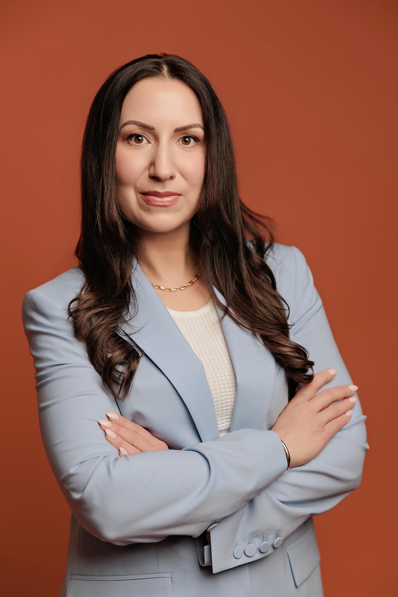 A woman with long dark hair wears a light blue blazer against a terracotta backdrop, showcasing the professional quality of this Calgary headshot studio.