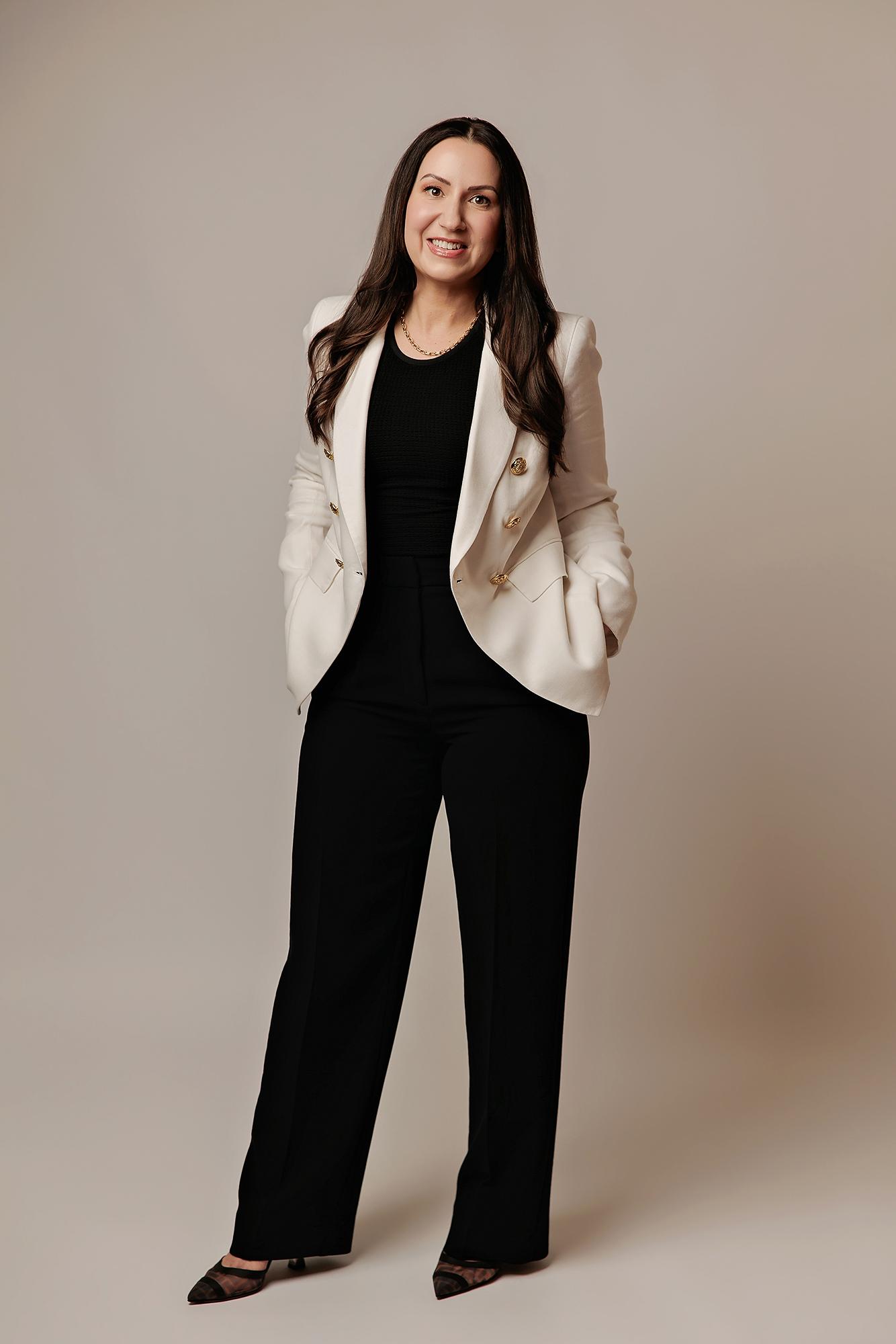 A smiling woman in a chic white blazer and black trousers stands in this Calgary photography studio, showcasing a professional look for personal branding.