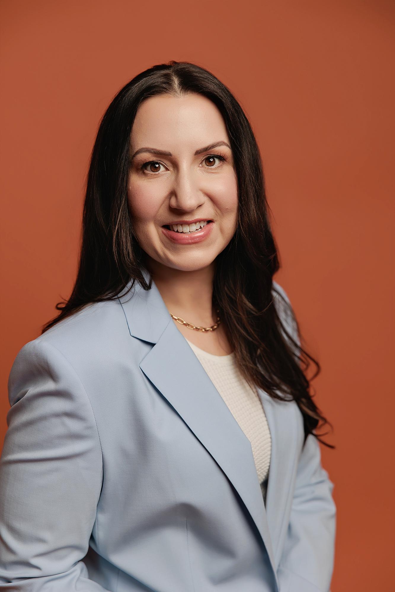 A smiling woman in a light blue blazer poses for a professional headshot at our Calgary photography studio, set against a warm terracotta backdrop.