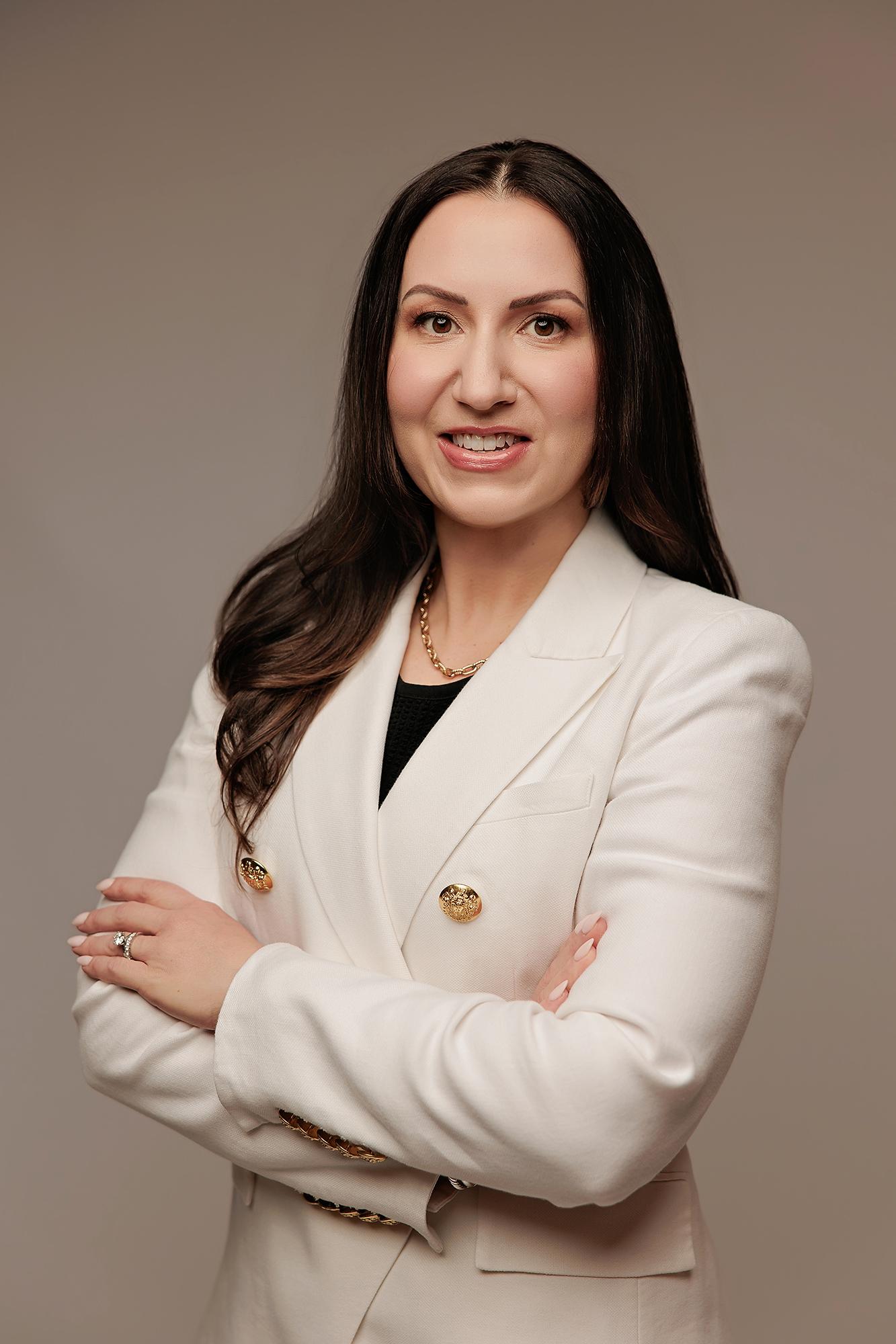 A professional woman with long dark hair smiles confidently in a white blazer, captured by a Calgary headshot photographer in a studio setting.