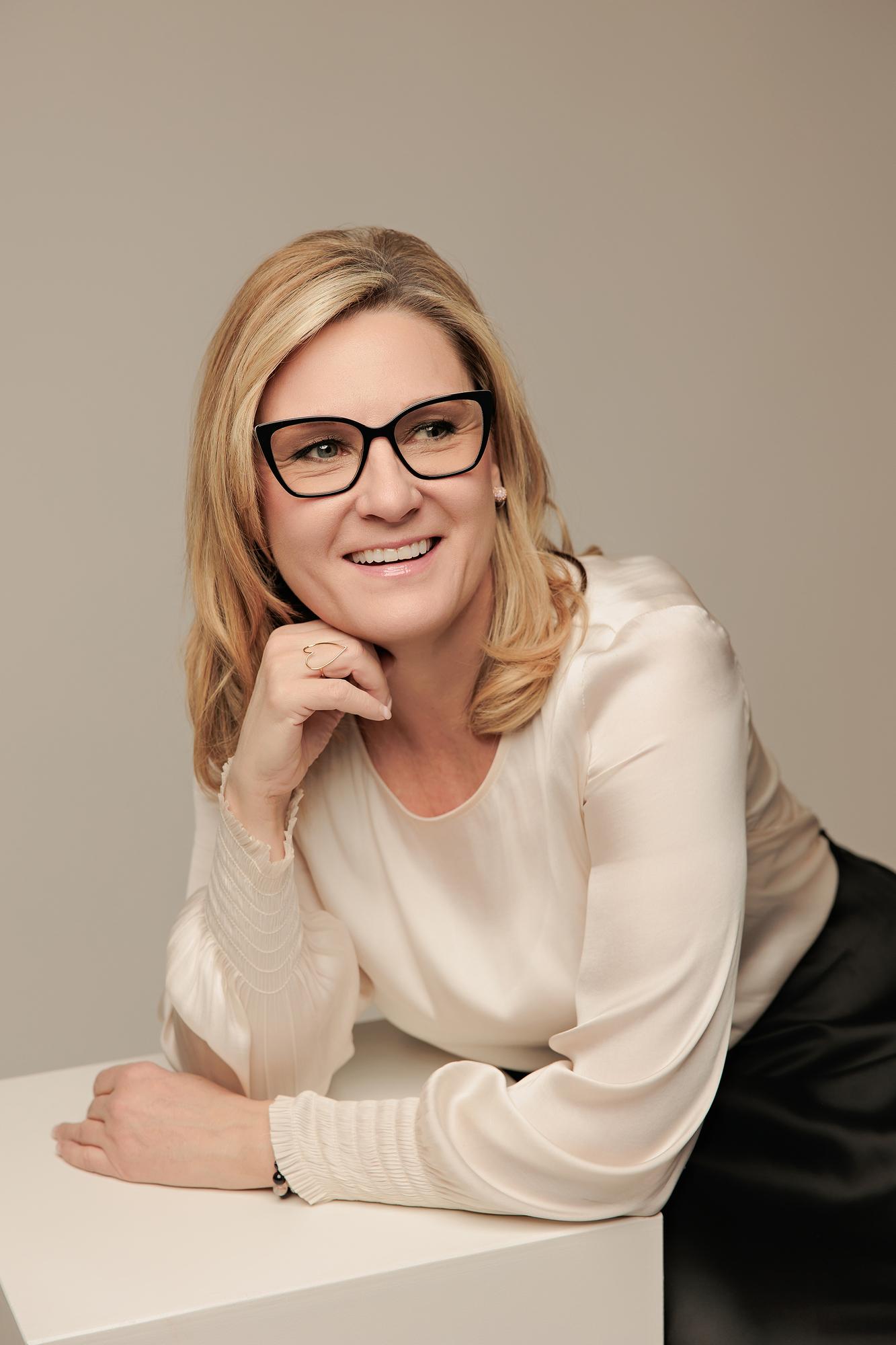 Warm professional headshot Calgary studio: Smiling woman with bold black-frame glasses in a cream silk blouse leaning on a white surface with her chin resting on her hand, taupe background
