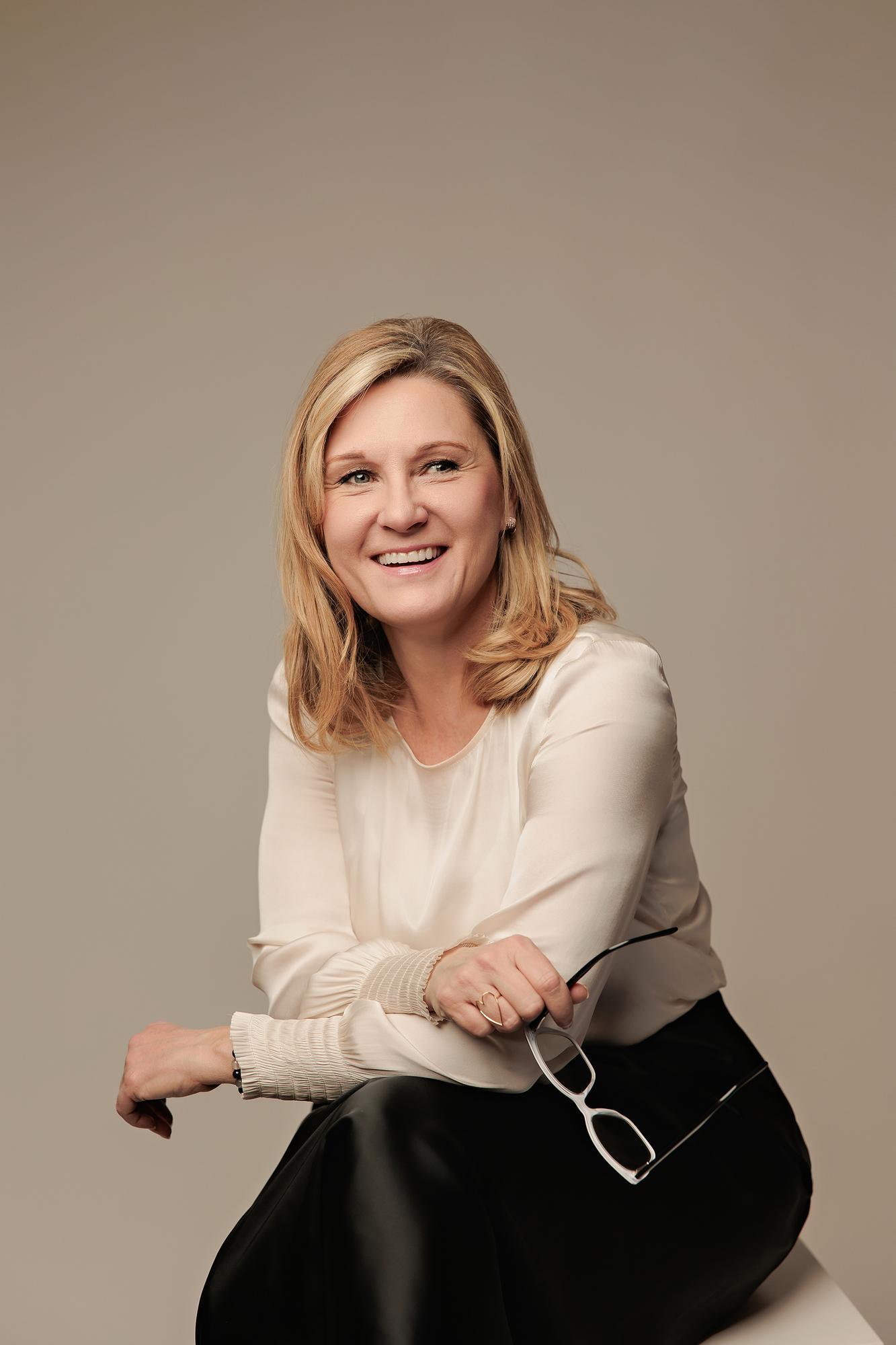 Professional headshot Calgary: Blonde woman in a cream silk blouse and black faux leather pants seated and leaning forward holding white glasses, warm taupe studio background