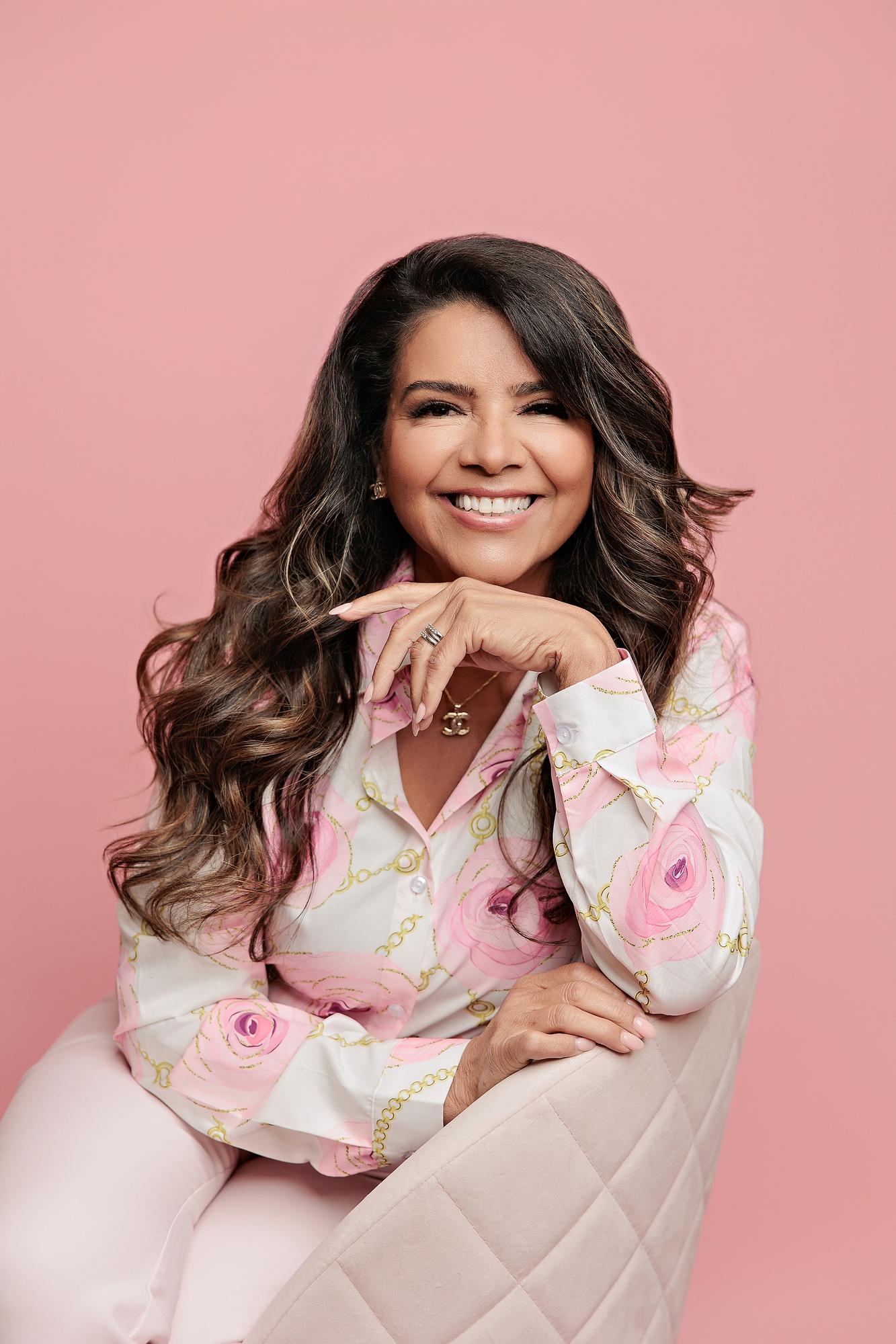 Personal branding photography Calgary: Glamorous businesswoman with long wavy hair seated on a quilted velvet chair with chin resting on hand, wearing a pink floral printed blouse against a pink studio background