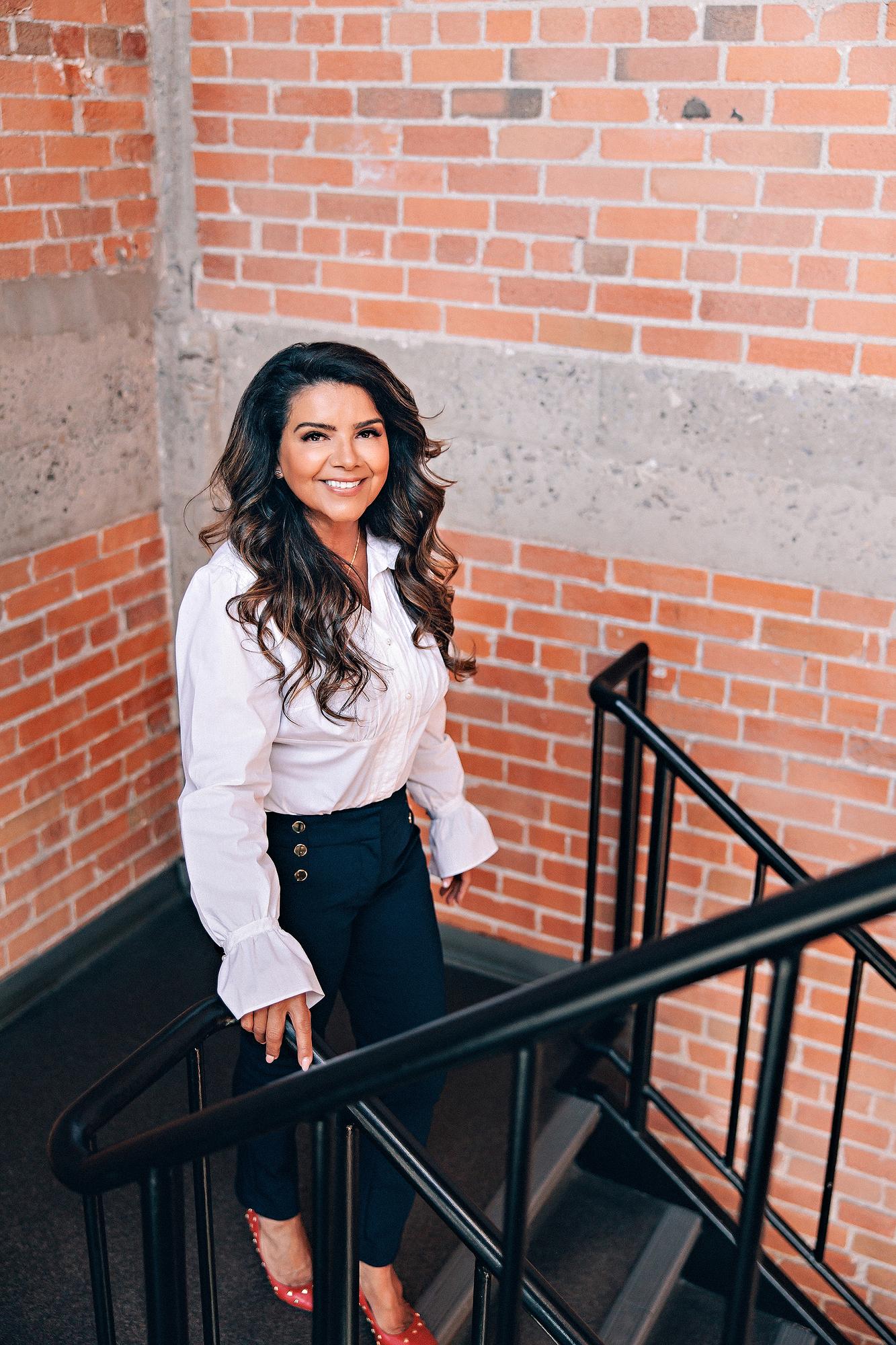 On-location personal branding headshot Calgary: Confident businesswoman in a white ruffled blouse and navy gold-button trousers smiling warmly on a brick staircase with black iron railings