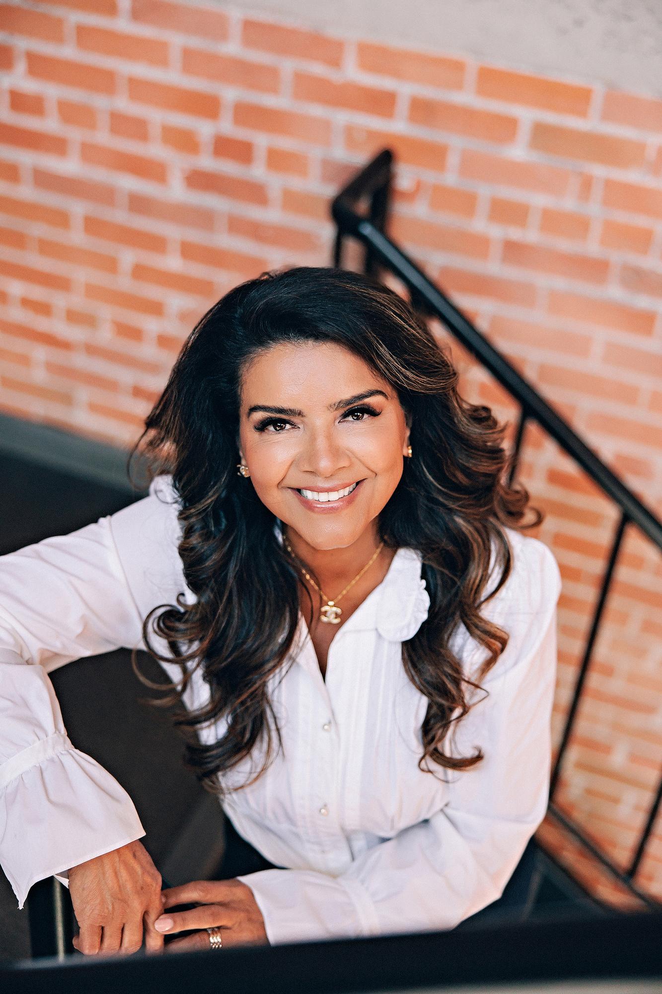 Close-up personal branding portrait Calgary: Warm close-up of a businesswoman in a white ruffled blouse leaning on black iron stair railing against an exposed brick wall