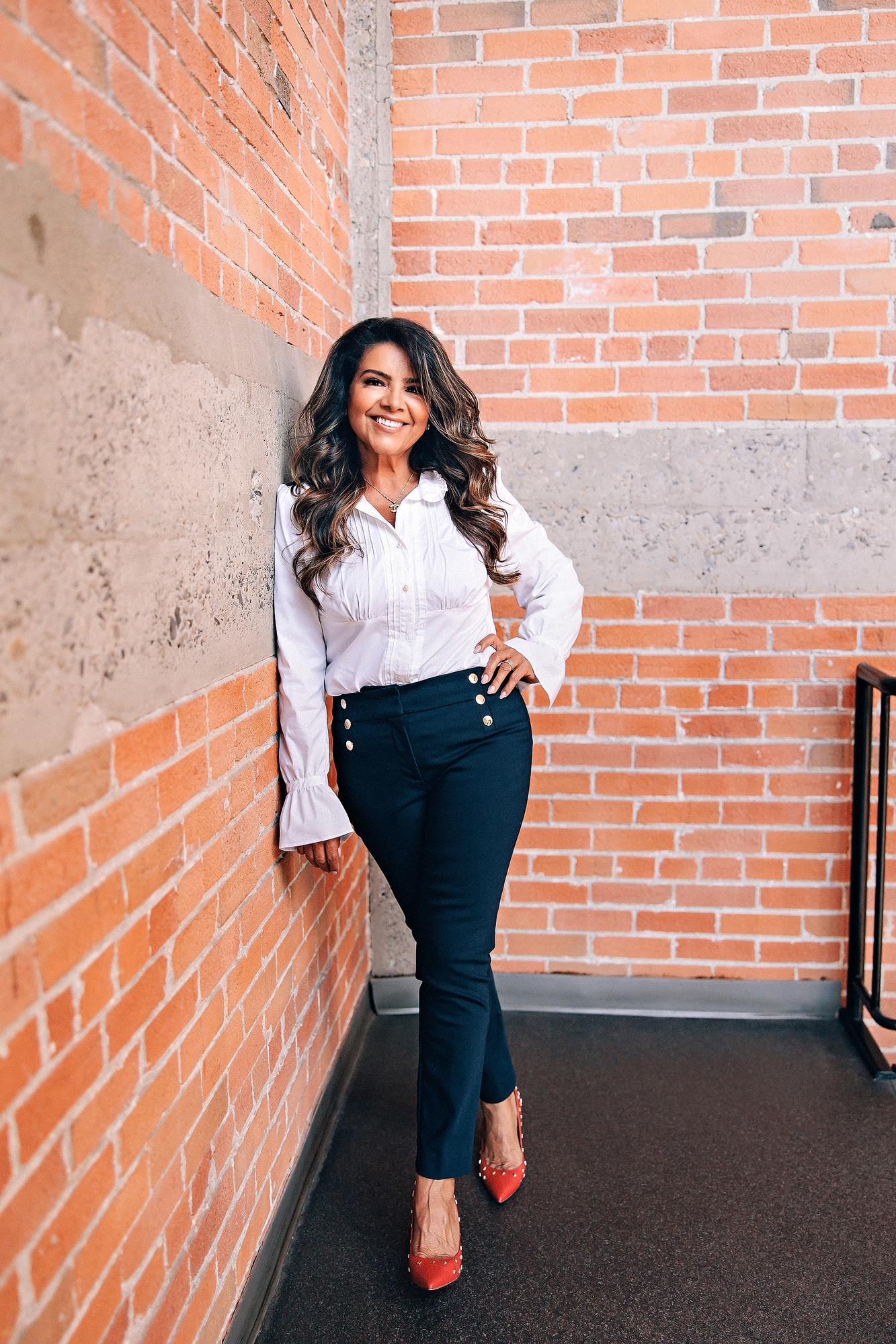 On-location personal branding photography Calgary: Full-length portrait of a confident professional woman in a white ruffled blouse and navy trousers leaning against an exposed brick wall with hand on hip and red studded heels