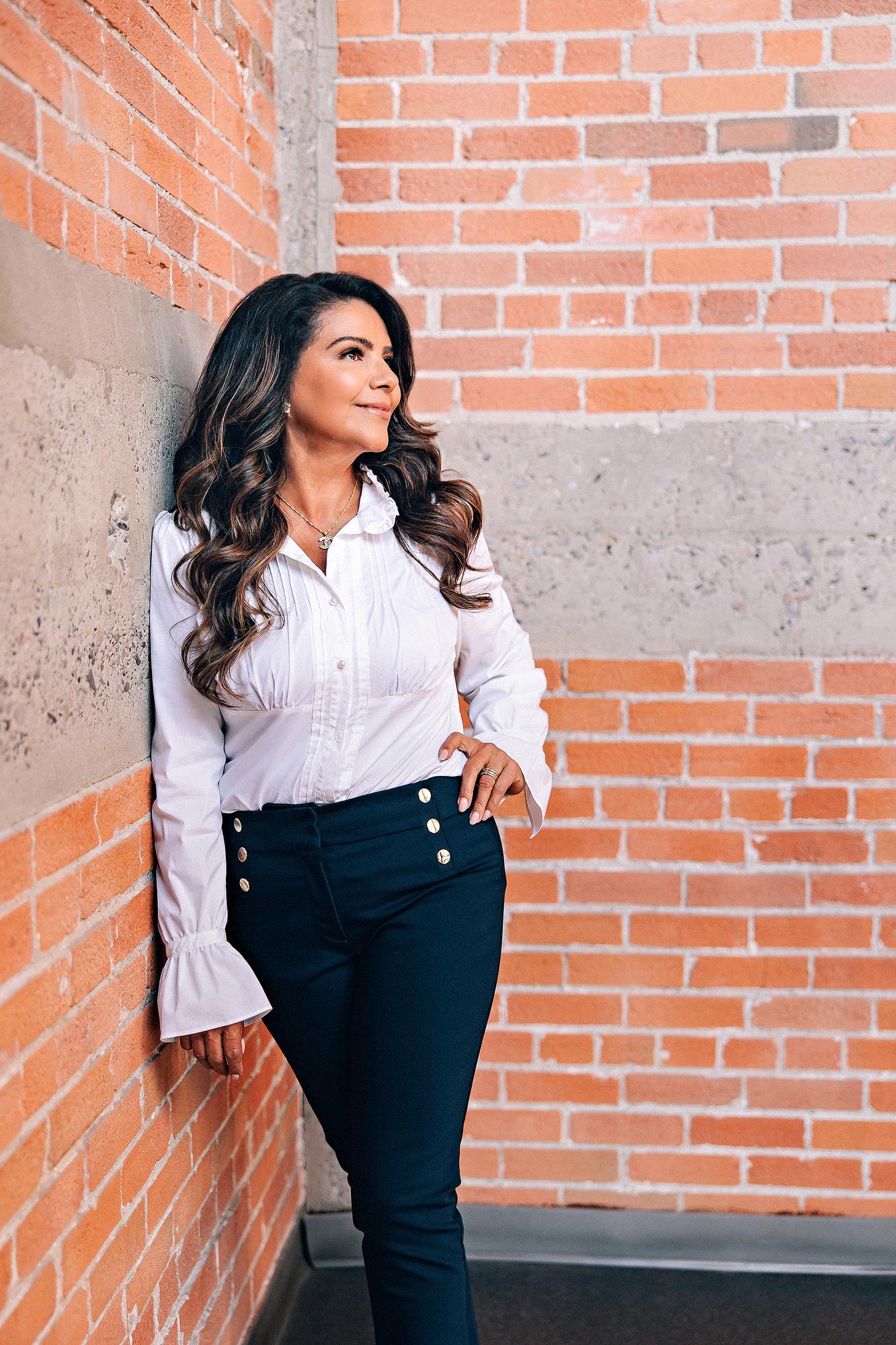 Environmental personal branding headshot Calgary: Poised businesswoman in a white ruffled blouse and navy gold-button trousers leaning against a brick wall and gazing upward with a serene expression