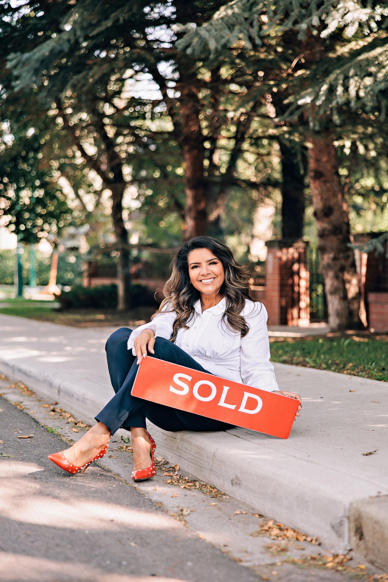 Real estate personal branding session Calgary: Joyful female realtor in a white ruffled blouse and navy trousers sitting on a curb holding a red Sold sign, surrounded by lush trees and red studded heels