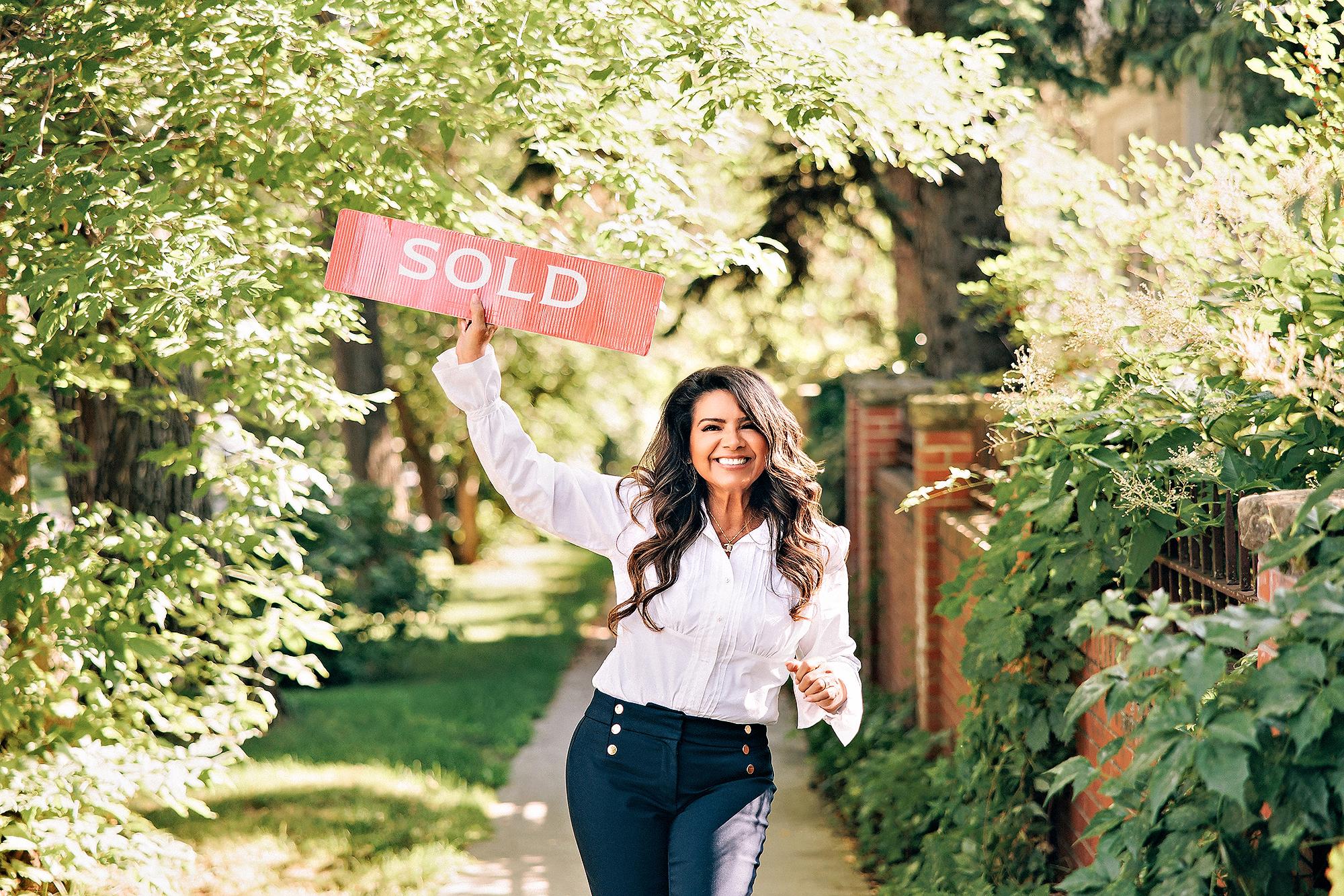 Outdoor personal branding photography Calgary: Energetic female realtor in a white ruffled blouse and navy trousers running down a tree-canopied garden path while holding a red Sold sign overhead