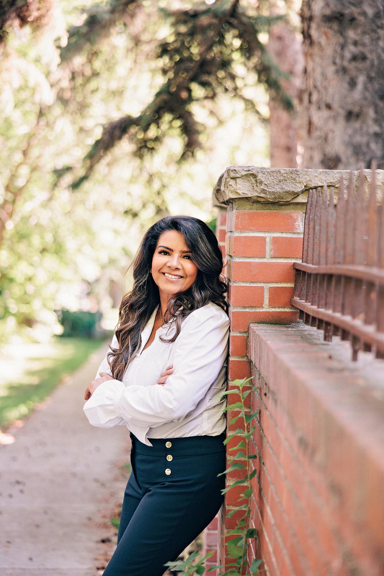 Outdoor personal branding headshot Calgary: Confident businesswoman in a white ruffled blouse and navy trousers leaning against a brick garden wall with arms crossed and a warm smile, lush trees in background