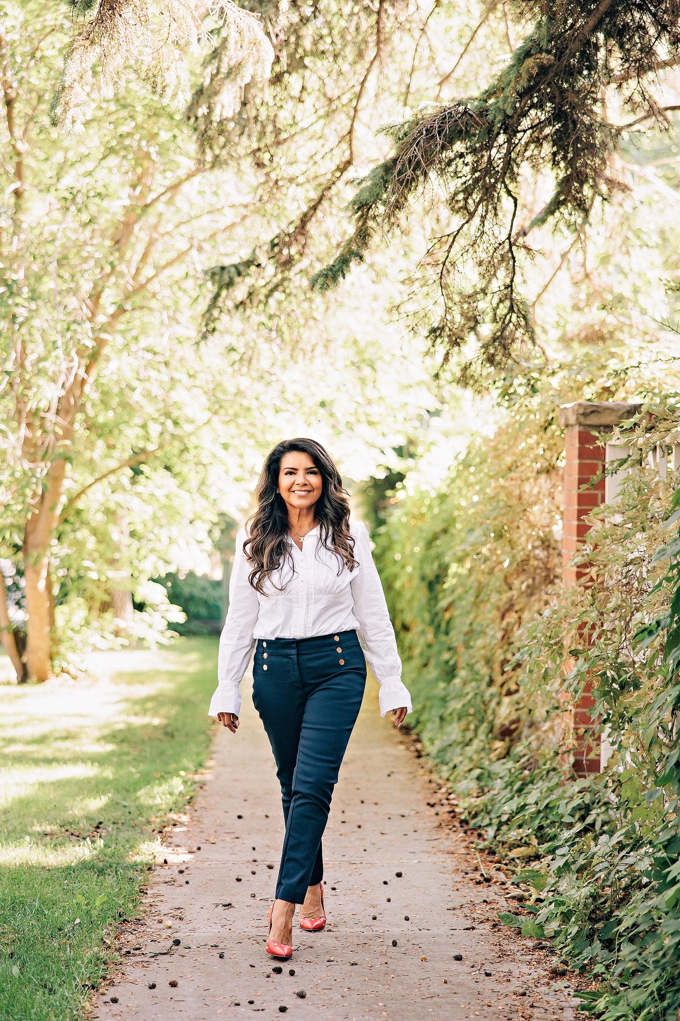 Lifestyle personal branding photography Calgary: Smiling professional woman in a white ruffled blouse and navy gold-button trousers walking toward camera along a dappled sunlit garden path with red studded heels
