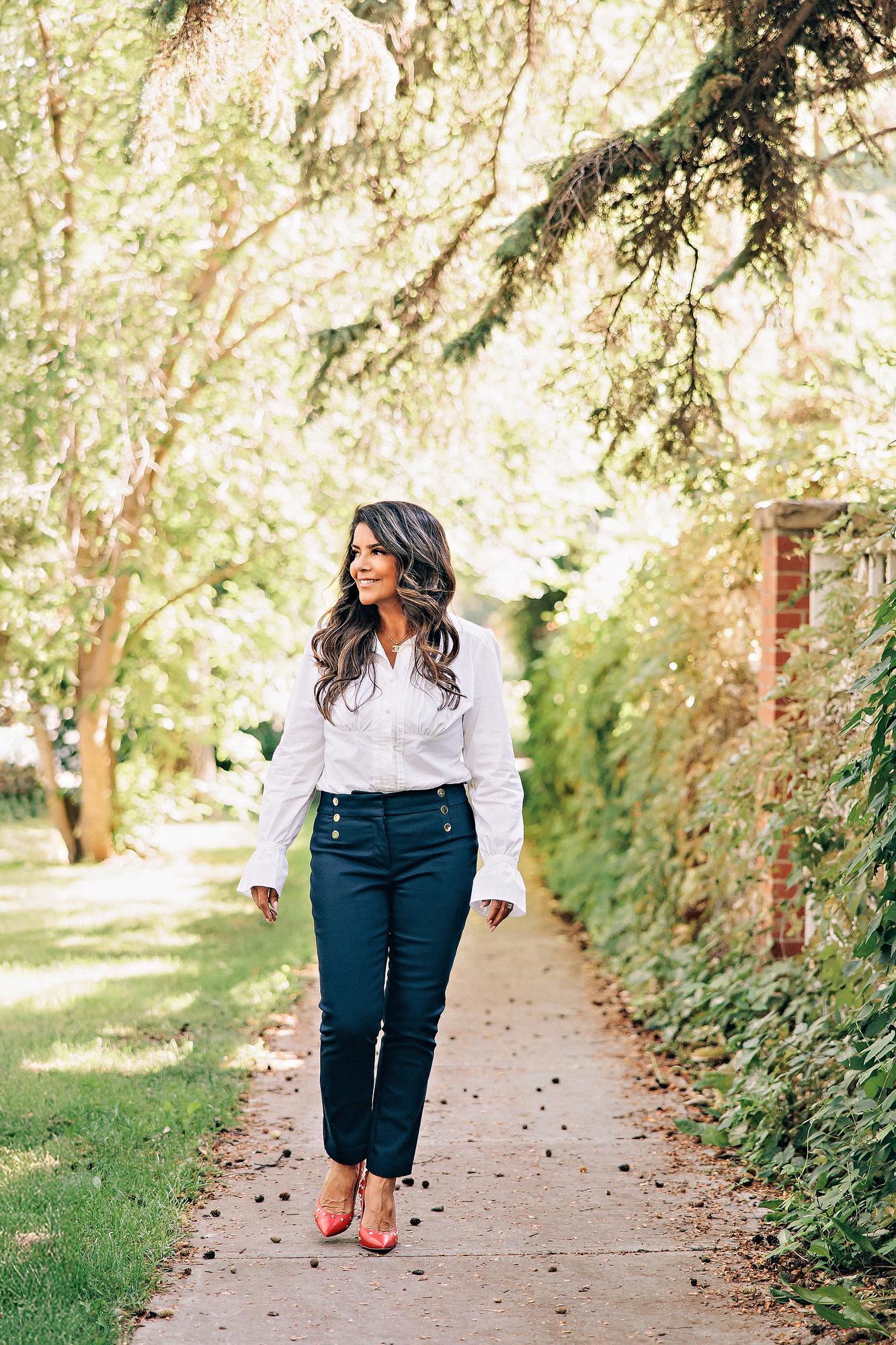 Outdoor personal branding session Calgary: Relaxed businesswoman in a white ruffled blouse and navy trousers strolling along a sunlit tree-lined path, glancing to the side with a natural smile and red heels