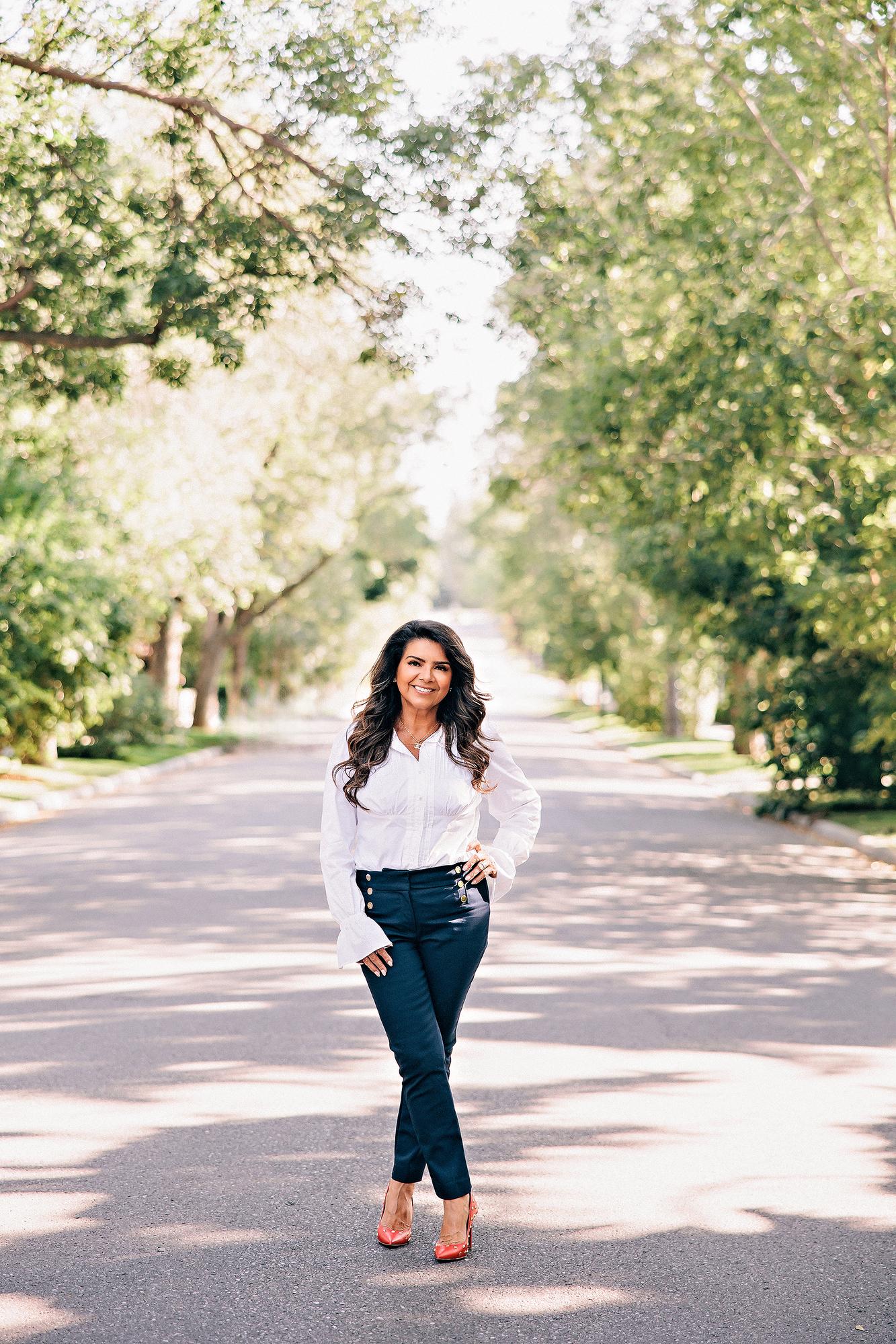 Full-length outdoor personal branding portrait Calgary: Poised female professional in a white ruffled blouse and navy gold-button trousers standing confidently on a tree-lined residential street with hand on hip and red studded heels