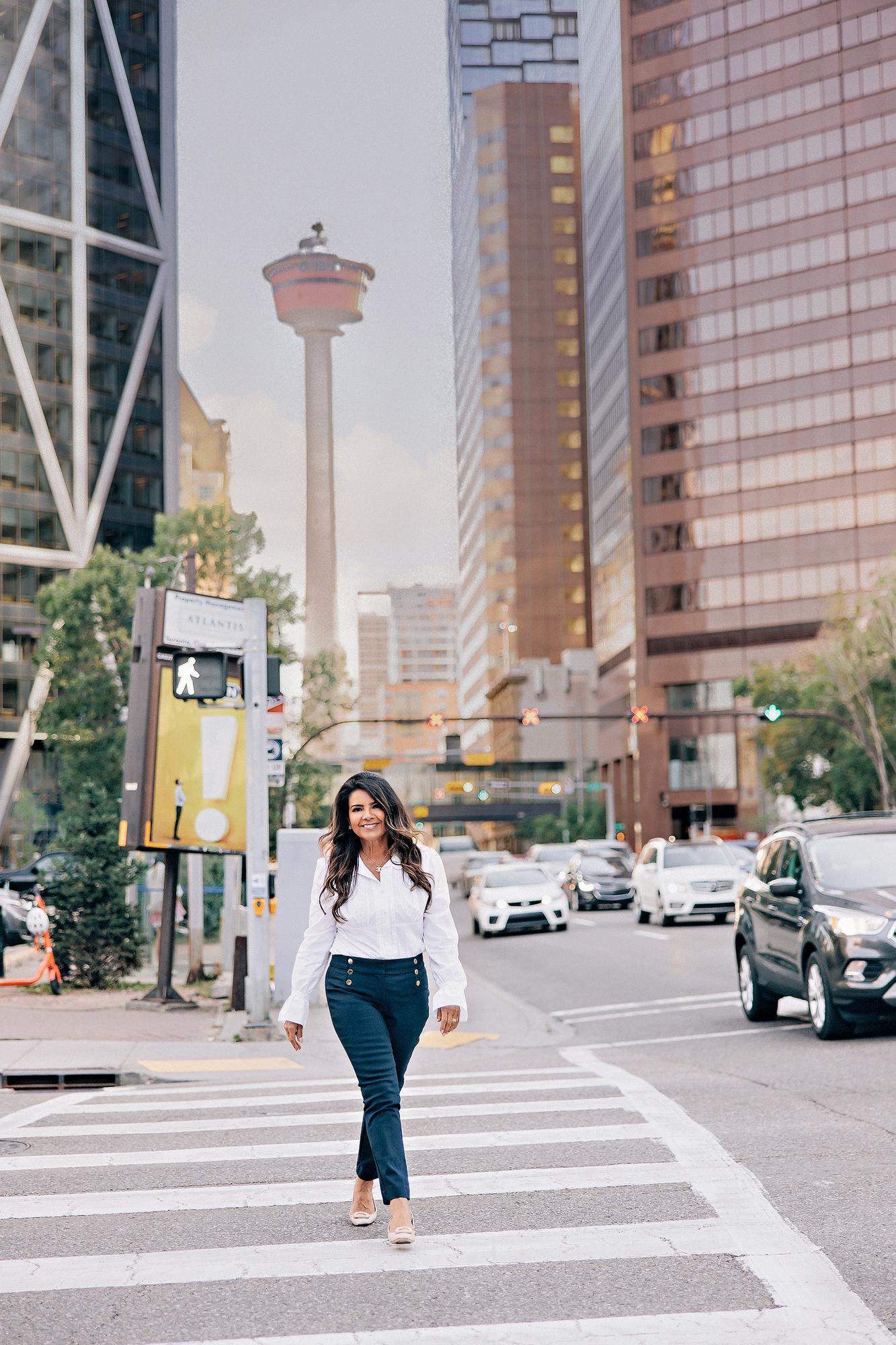 Urban personal branding headshot Calgary downtown: Smiling professional woman in a white ruffled blouse and navy gold-button trousers walking across a zebra crossing with the Calgary Tower rising between downtown skyscrapers in the background