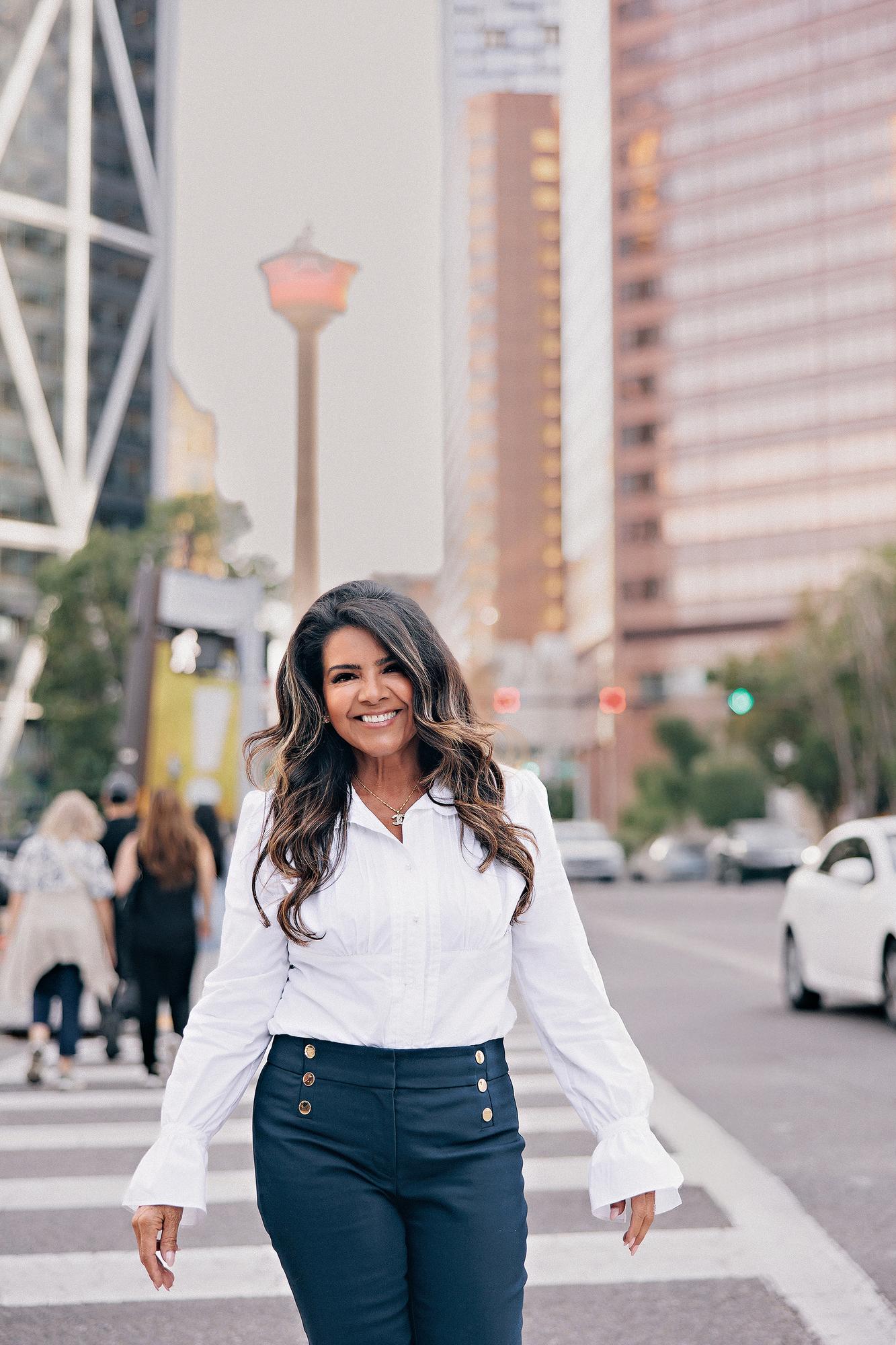 Urban personal branding photography Calgary downtown: Smiling businesswoman in a white ruffled blouse and navy gold-button trousers walking confidently across a busy crosswalk with the Calgary Tower visible in the background