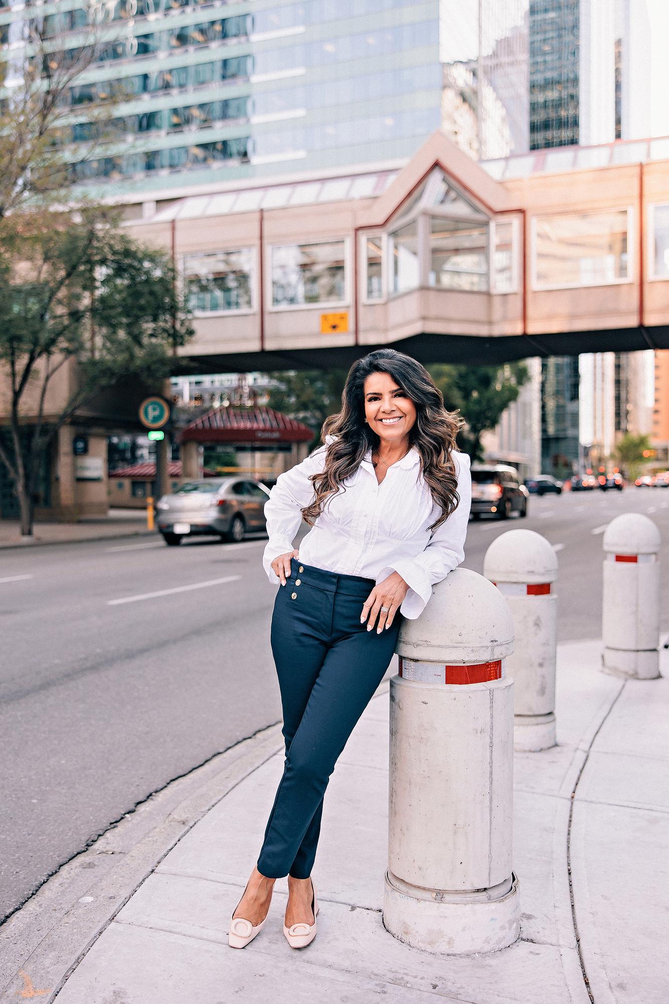 Downtown Calgary personal branding headshot: Confident female professional in a white ruffled blouse and navy trousers leaning casually against a bollard on a city sidewalk with downtown office towers in the background