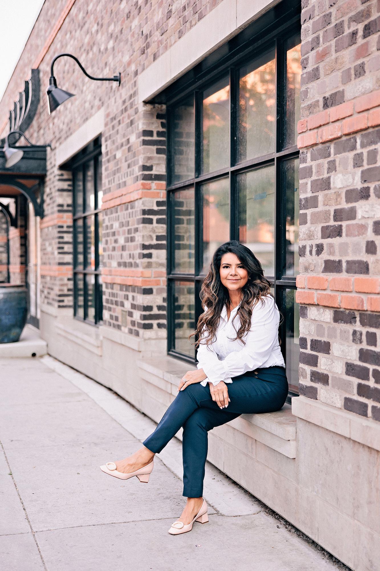 On-location personal branding photography Calgary: Relaxed businesswoman in a white ruffled blouse and navy trousers seated on a stone window ledge outside a brick building with black-framed industrial windows