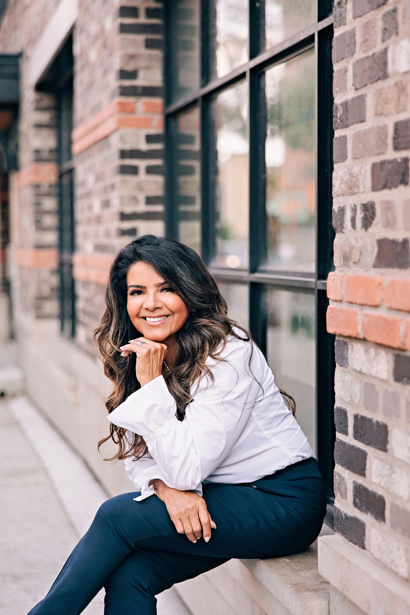 Outdoor personal branding headshot Calgary: Approachable female entrepreneur in a white ruffled blouse and navy trousers seated on a stone ledge with chin resting on hand and a warm smile, exposed brick and black-framed windows behind her
