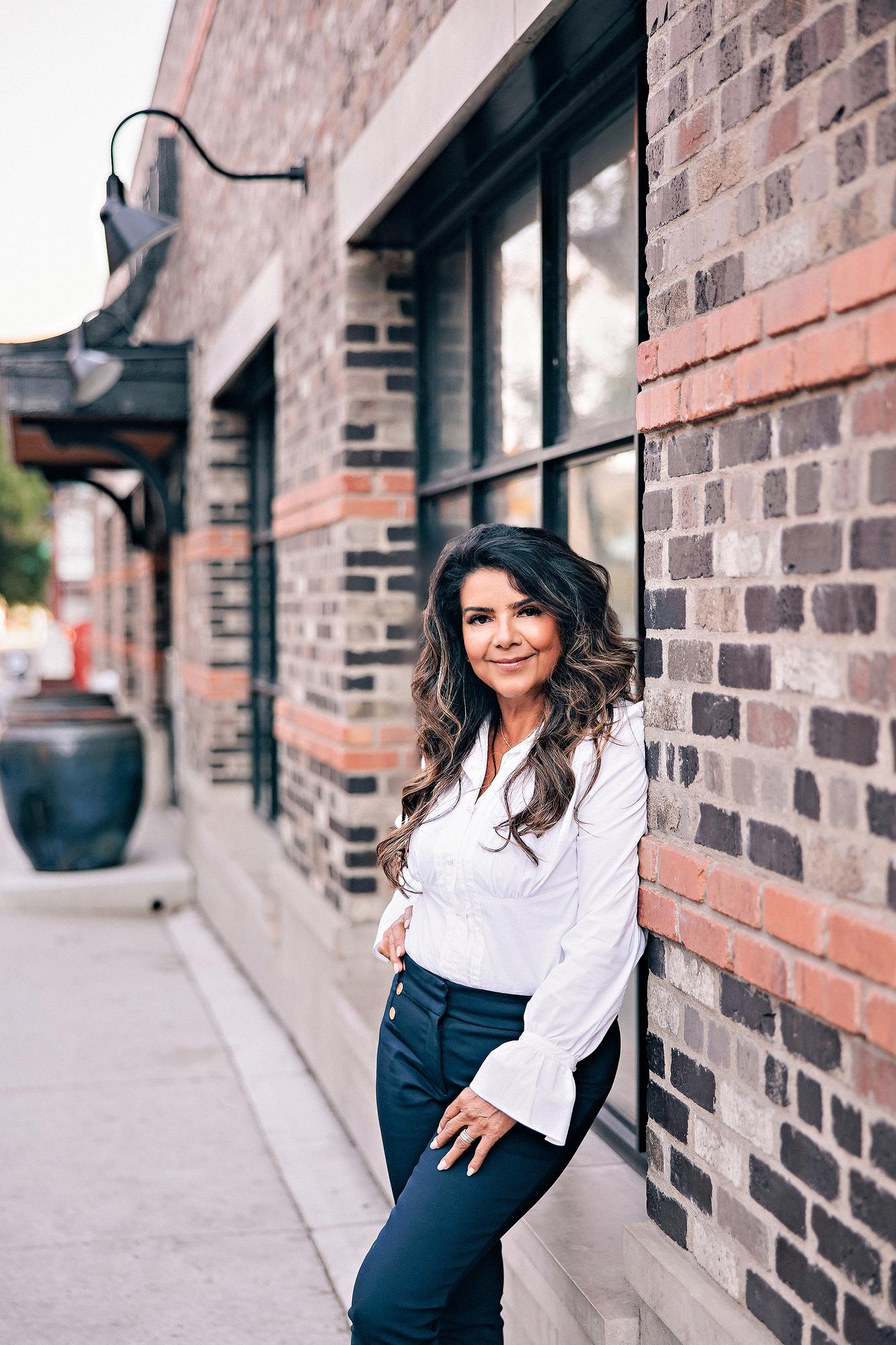 On-location personal branding session Calgary: Poised businesswoman in a white ruffled blouse and navy gold-button trousers leaning against an exposed brick wall beside black-framed industrial windows on a Calgary streetscape