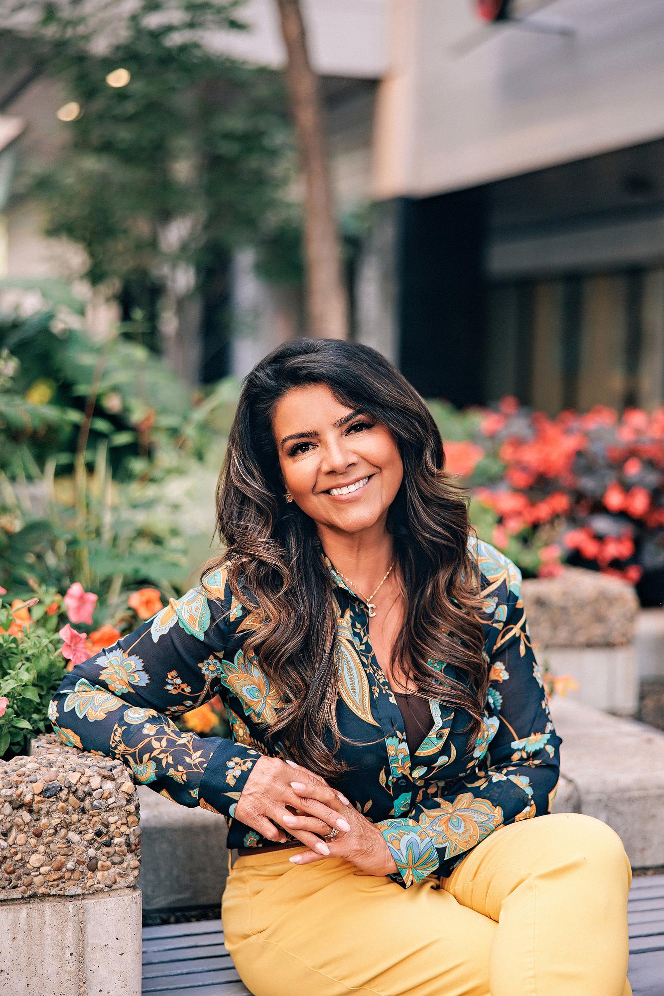 Lifestyle personal branding photography Calgary: Warm and approachable businesswoman in a dark floral paisley blouse and mustard yellow trousers seated on a bench with hands clasped, colourful flower gardens in the background