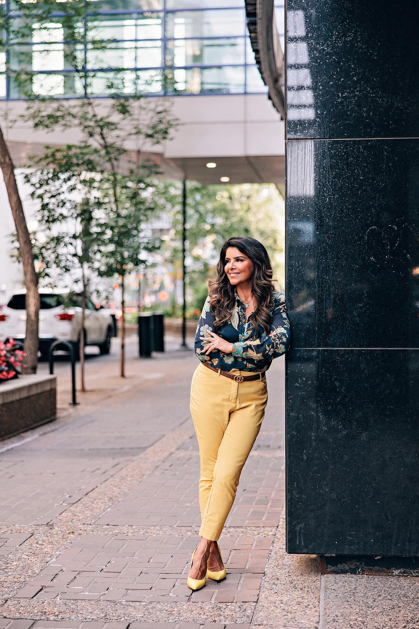 Urban personal branding headshot Calgary: Confident female entrepreneur in a dark floral paisley blouse and mustard yellow trousers leaning against a black granite pillar on a downtown Calgary sidewalk, glancing away with a soft smile