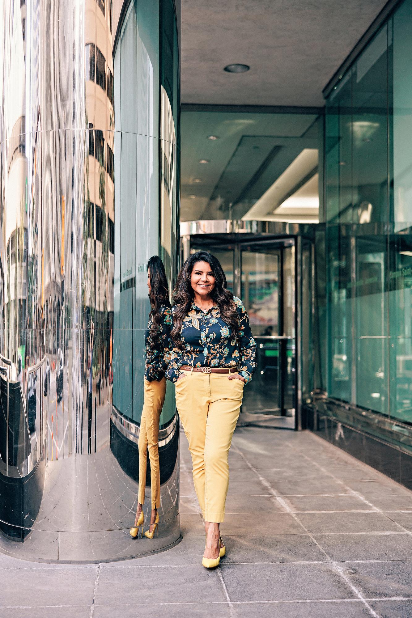 Downtown Calgary personal branding photography: Stylish businesswoman in a dark floral paisley blouse and mustard yellow trousers with matching heels standing beside a mirrored column at a Calgary office tower entrance, her reflection visible