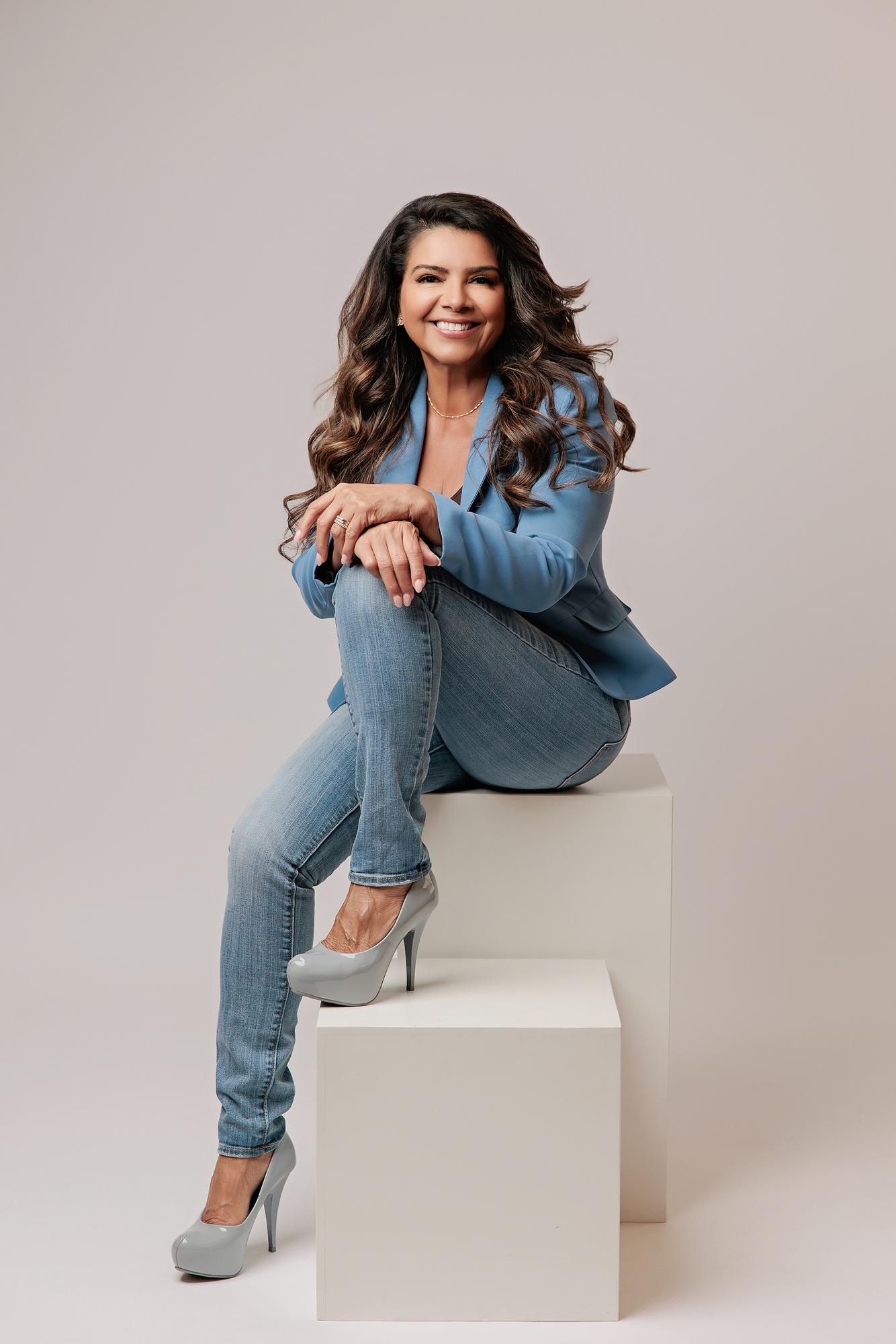 Personal branding photography Calgary: Professional woman in a steel blue blazer and jeans seated on a white cube working on a MacBook laptop, neutral grey studio background