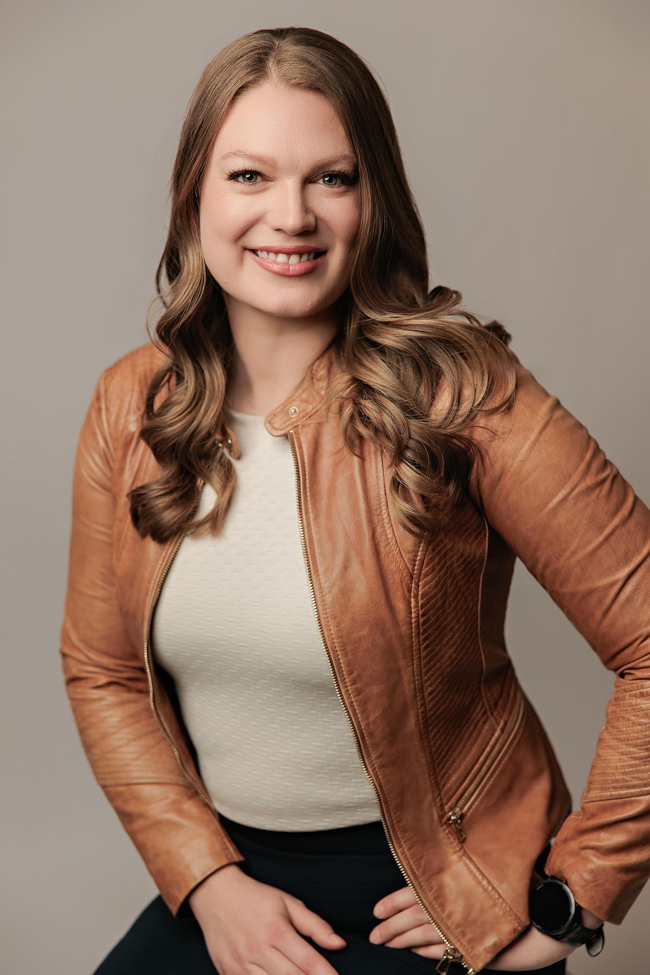 Professional headshot Calgary studio: Confident young woman with long wavy blonde-highlighted hair in a tan leather moto jacket and cream top standing with hands on hips and a bright smile against a warm grey background