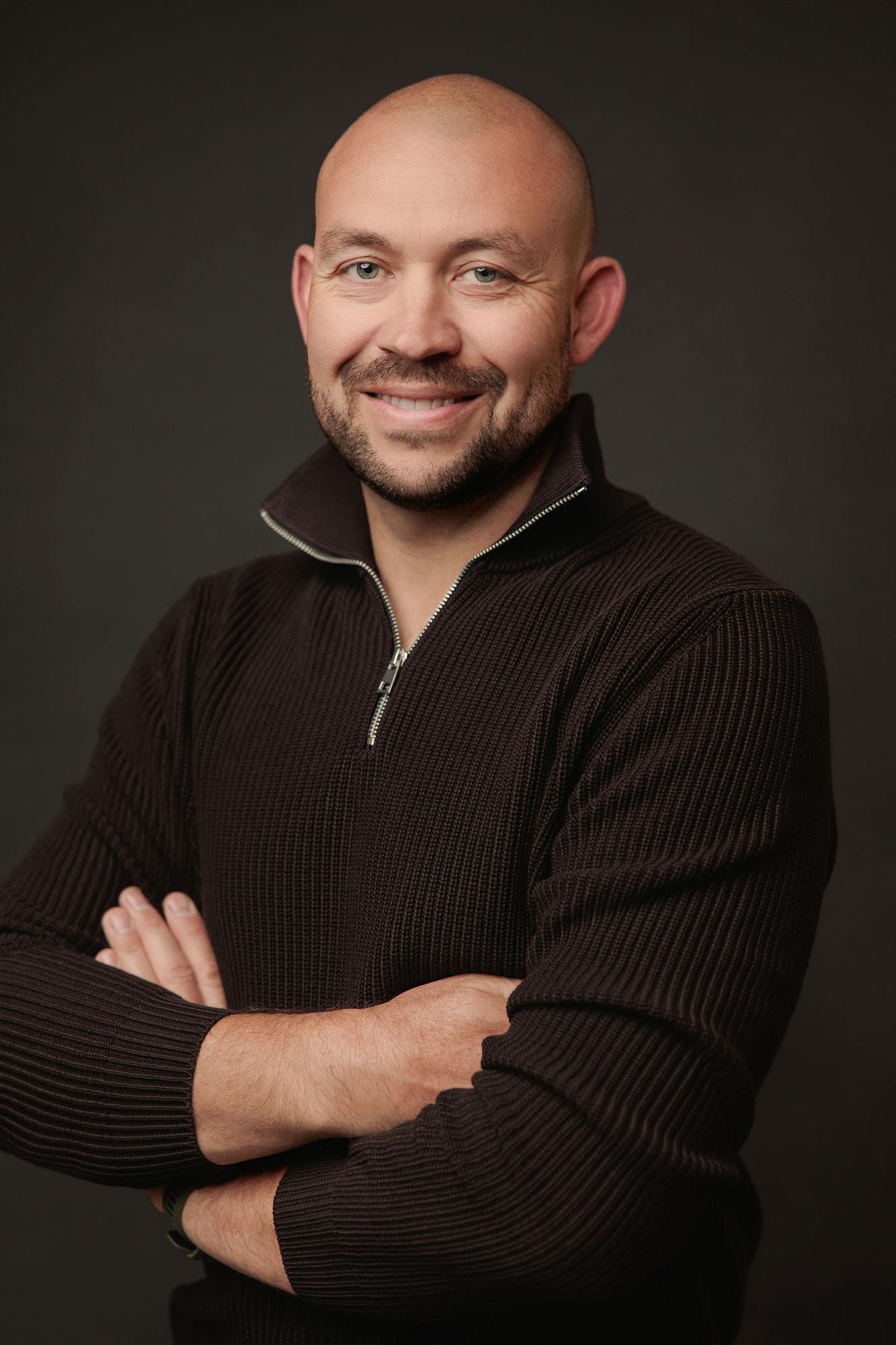 A smiling man in a dark knit sweater poses with arms crossed for this professional headshot captured at a Calgary photography studio.
