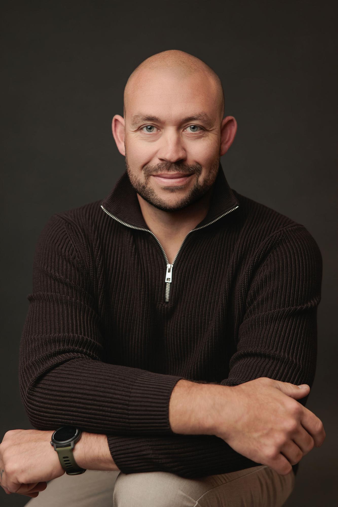 A bald man with a friendly smile wearing a dark zip-neck sweater poses for a professional headshot at our Calgary photography studio, studio lit.
