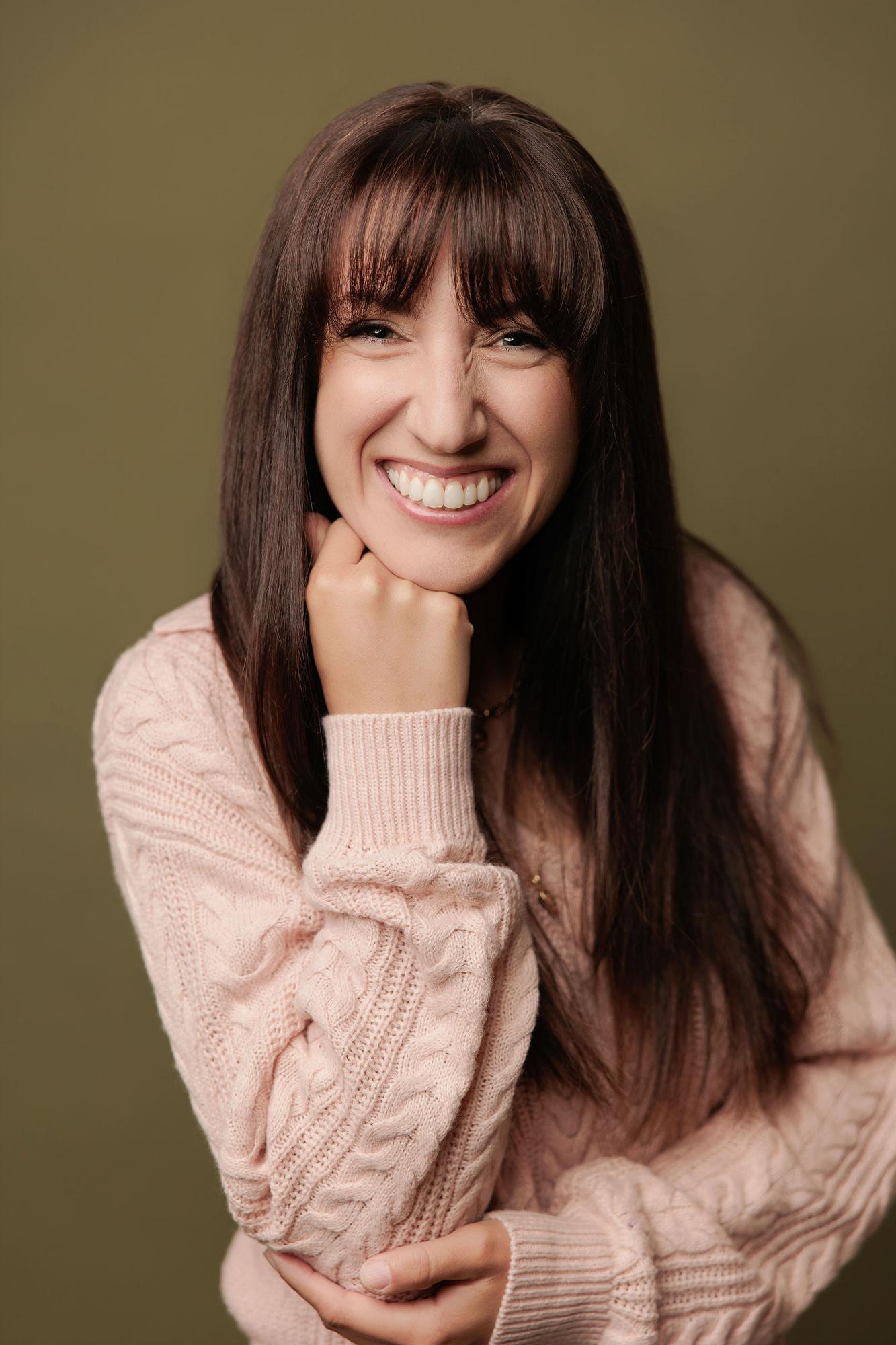 A woman with long dark hair and a pink sweater smiles warmly at our Calgary photography studio, showcasing the approachable style of our headshots.