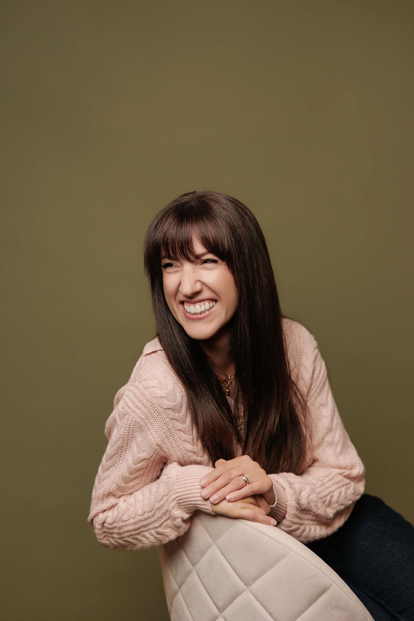 A woman with long dark hair laughs in a pink cable-knit sweater, captured by a Calgary headshot photographer in a studio with a warm olive backdrop.