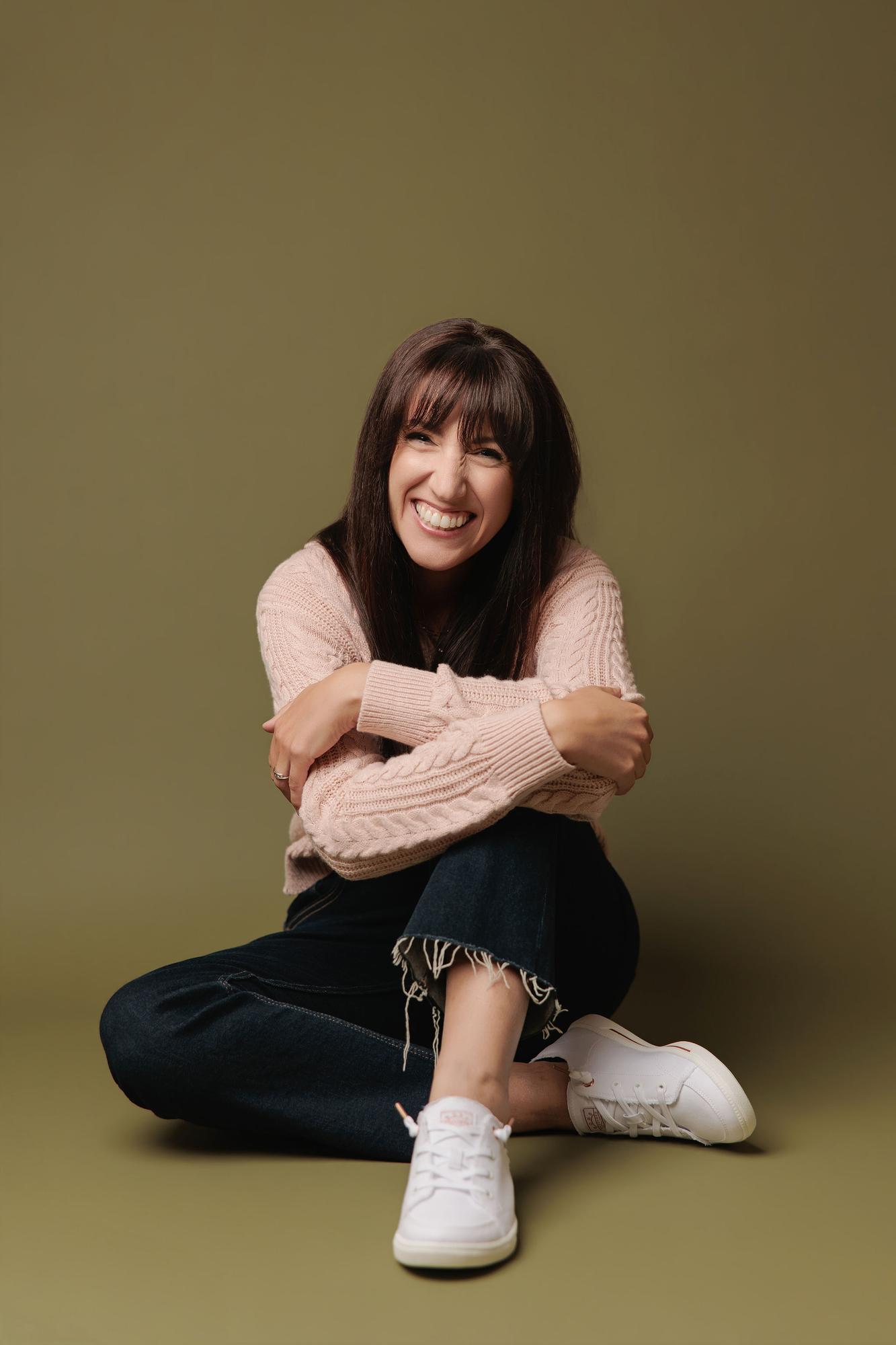 A woman with bangs laughs while sitting on the floor in a cozy pink sweater, showcasing the relaxed personal branding style of our Calgary studio.