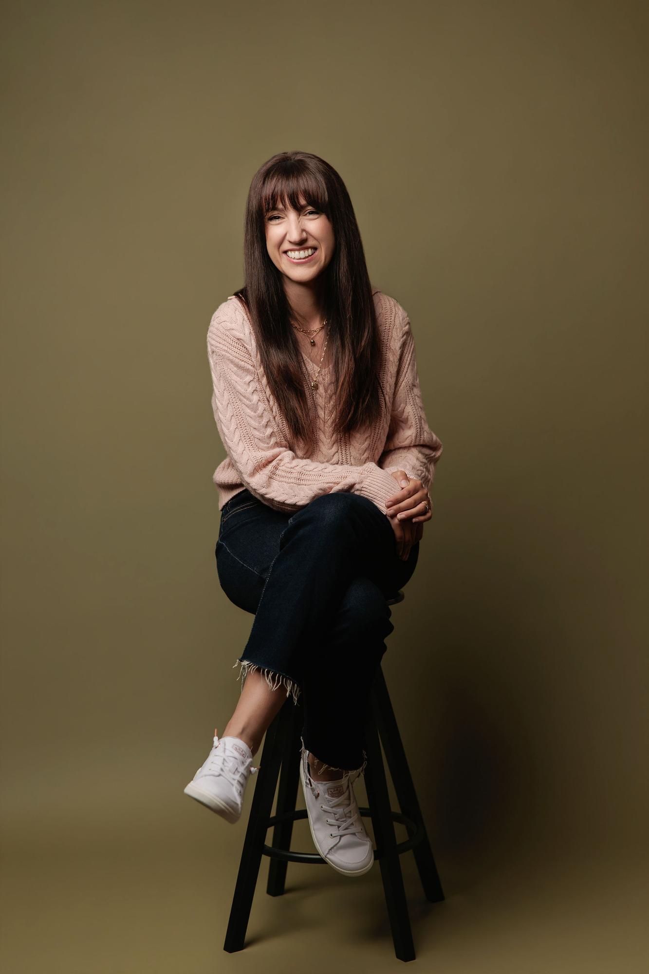 A smiling woman in a pink sweater sits on a stool against an olive backdrop, showcasing the approachable style of this Calgary headshot photographer.