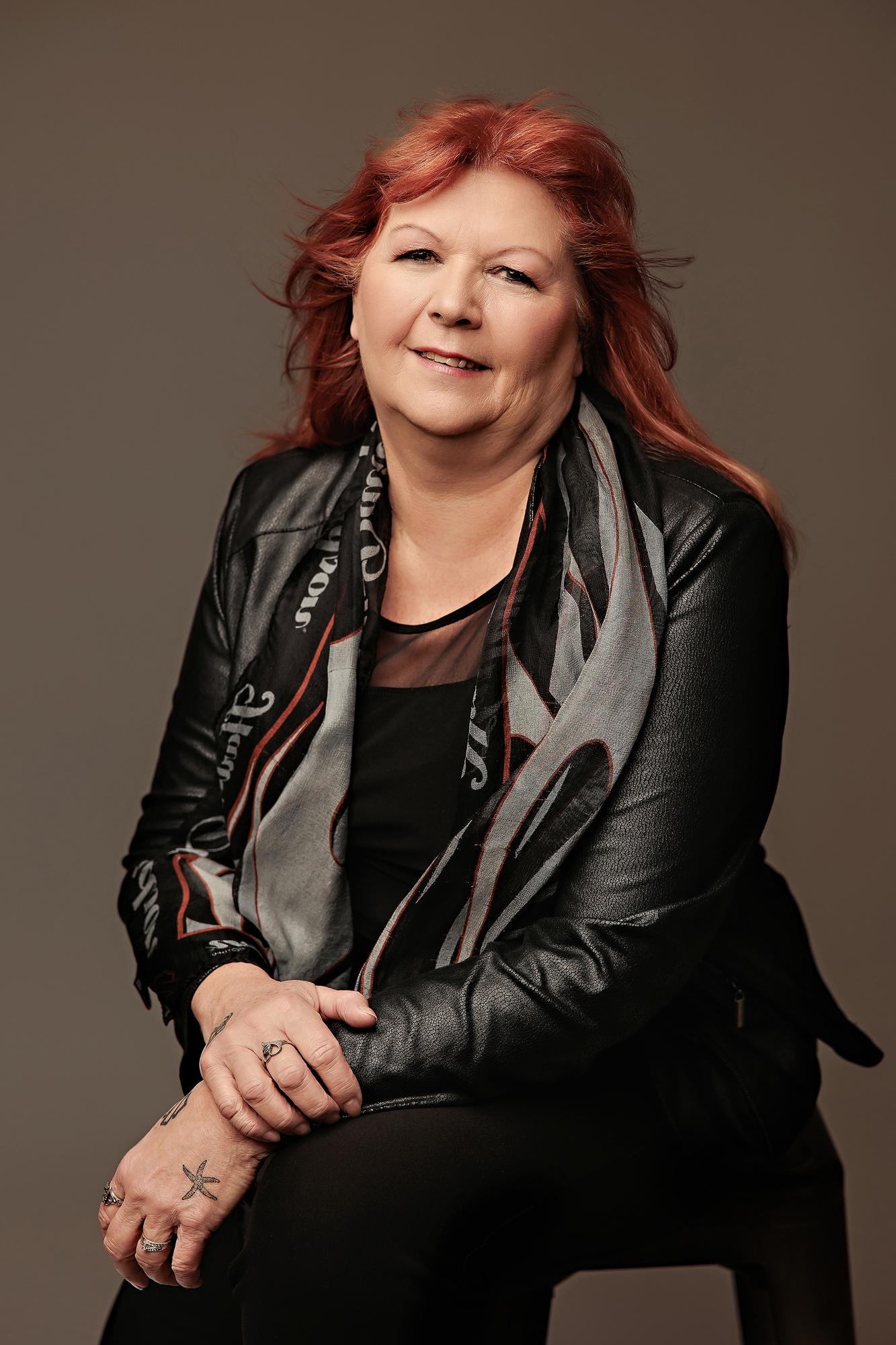 Professional headshot Calgary studio: Edgy female professional with vibrant red hair in a black leather jacket and graphic grey scarf seated with hands clasped on a dark stool against a warm taupe background
