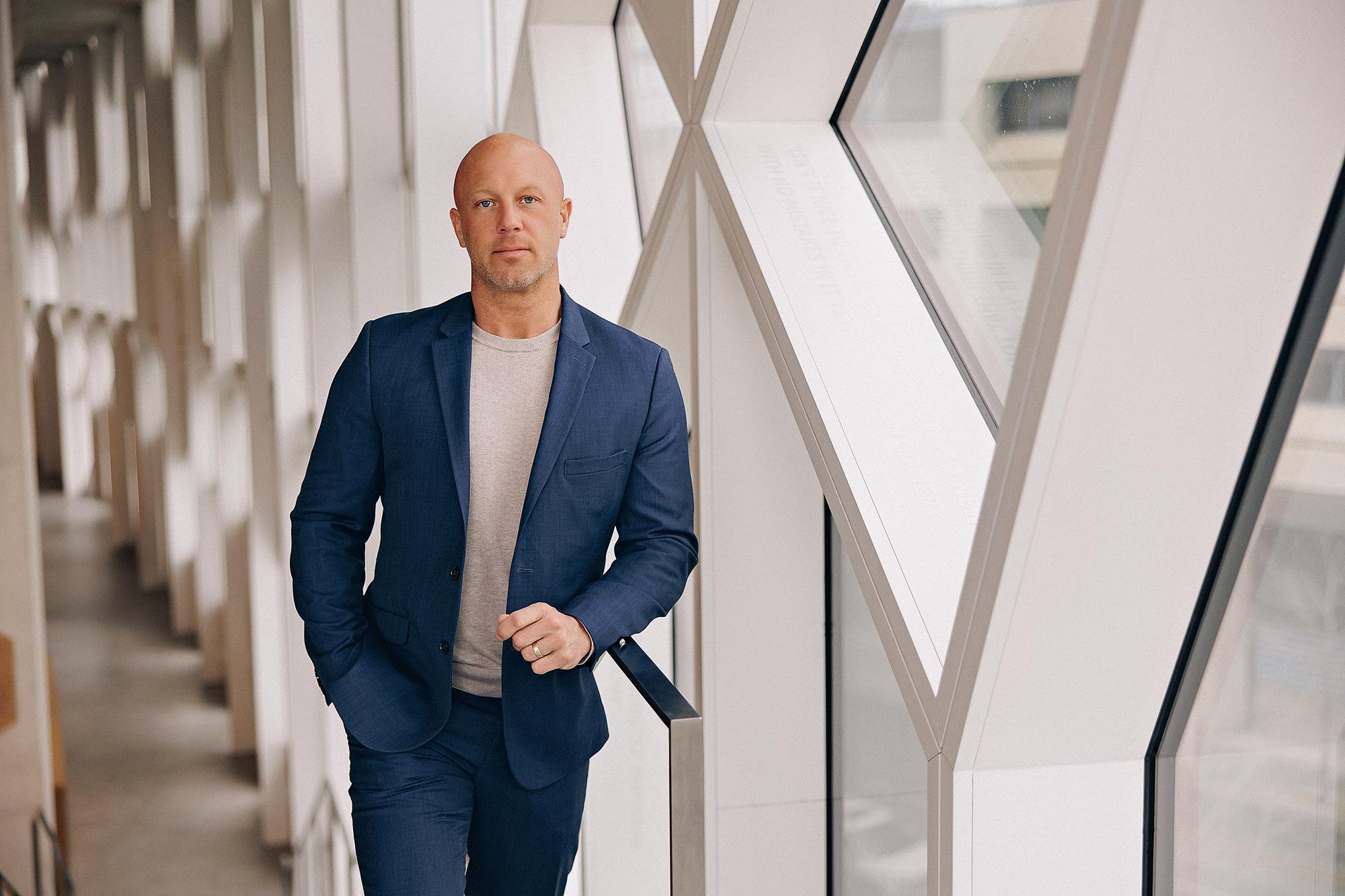 On-location personal branding photography Calgary: Confident professional man in a navy blue blazer and grey t-shirt leaning against a railing in a dramatic angular glass and steel corridor inside the Bow Tower