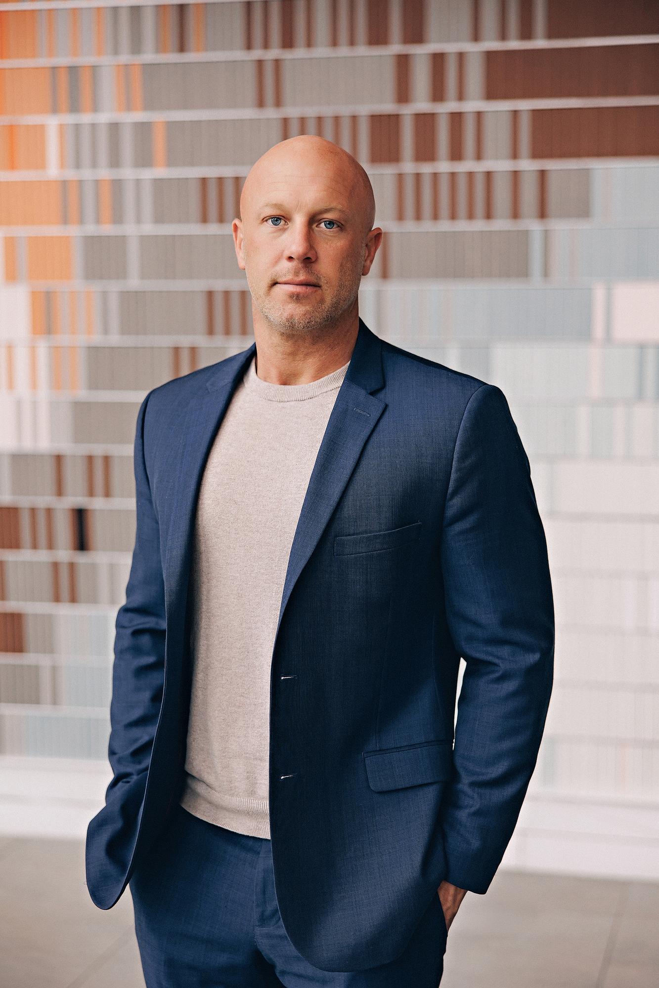 Personal branding headshot Calgary: Authoritative male executive in a navy blue blazer and grey t-shirt standing with hands in pockets in front of a vibrant multicolour striped art wall inside a Calgary office tower