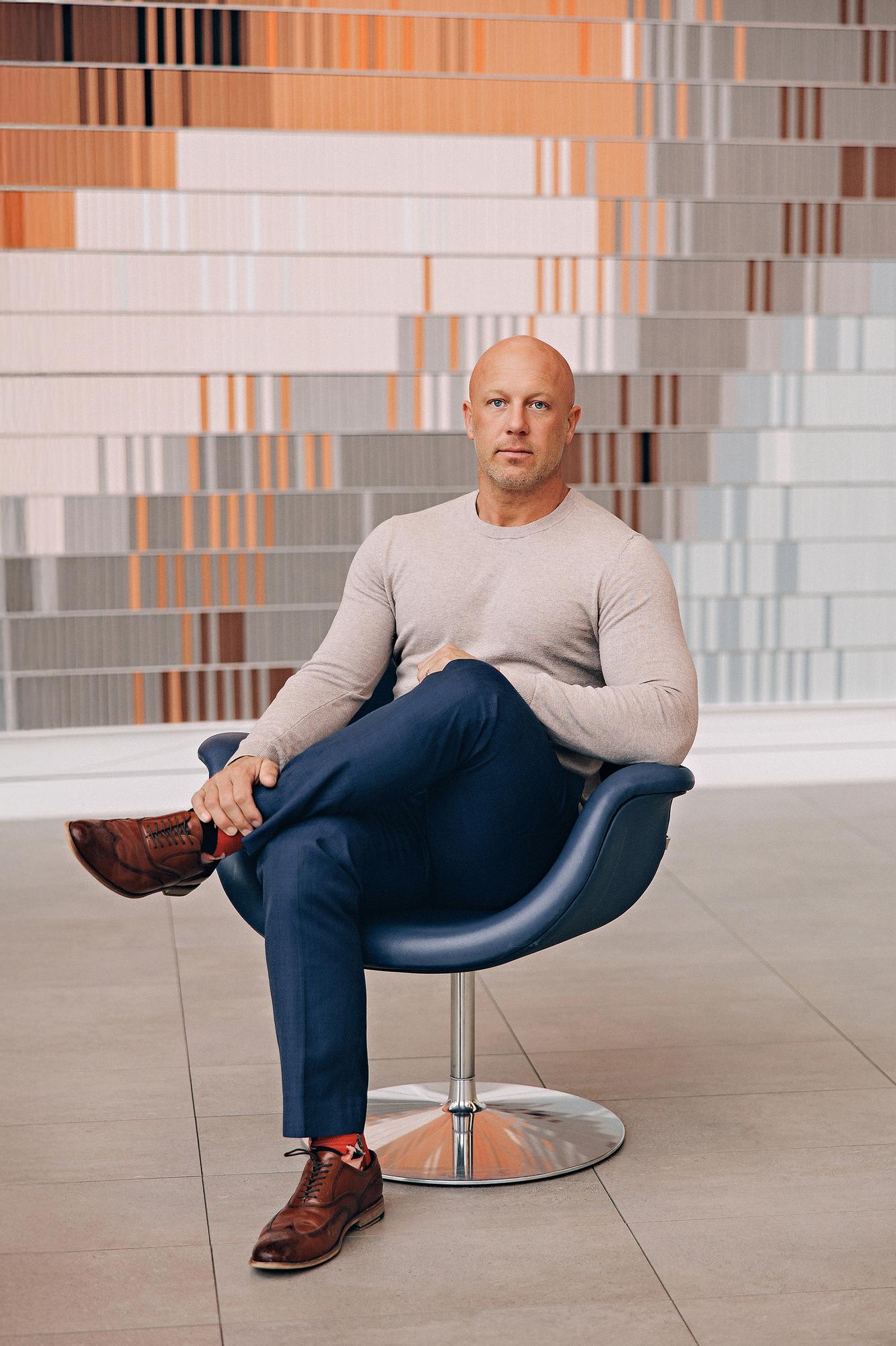On-location personal branding photography Calgary: Relaxed professional man in a grey sweater and navy trousers seated with one leg crossed in a sculptural blue leather lounge chair in front of a colourful striped art wall