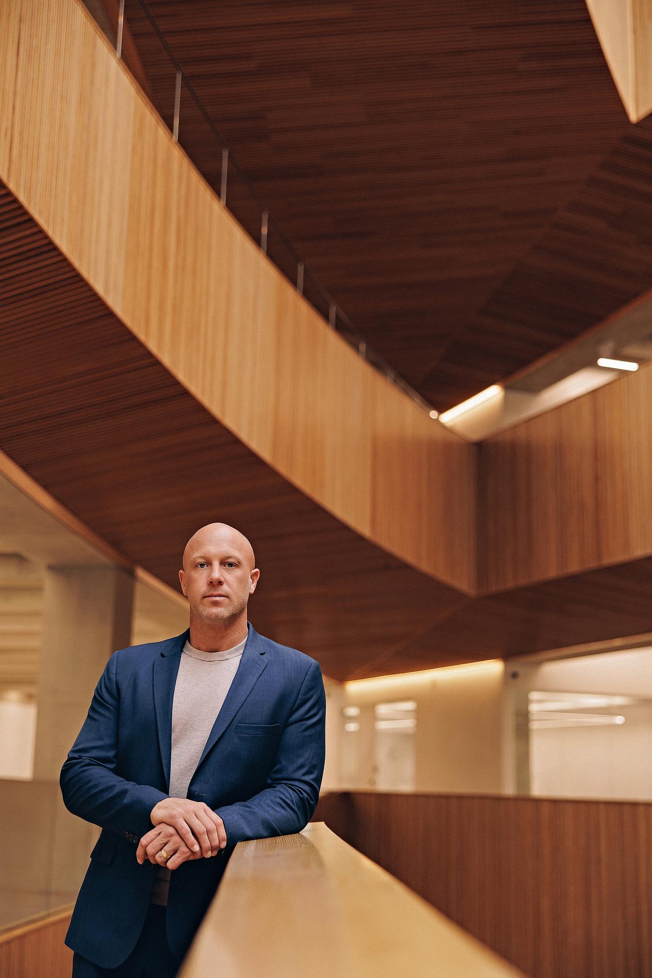 Environmental personal branding photography Calgary: Brooding male professional in a navy blue blazer and grey t-shirt leaning on a wood railing inside a dramatic curved wood-panelled atrium with warm ambient lighting