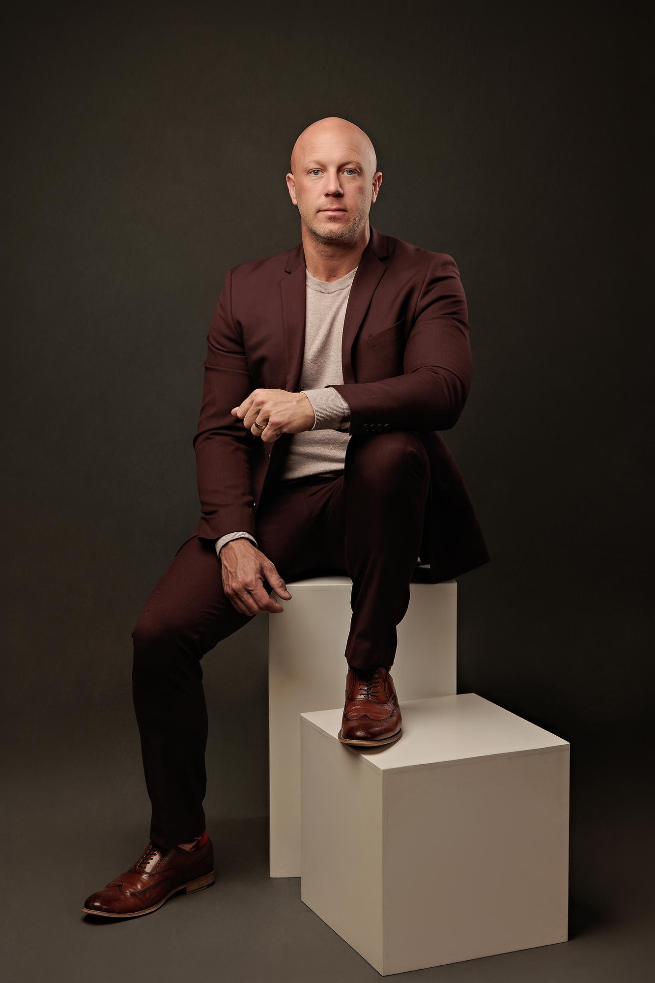 Professional headshots Calgary studio: Commanding male executive in a deep burgundy blazer and grey t-shirt seated on stacked white studio cubes with one hand resting on his knee, dark charcoal background