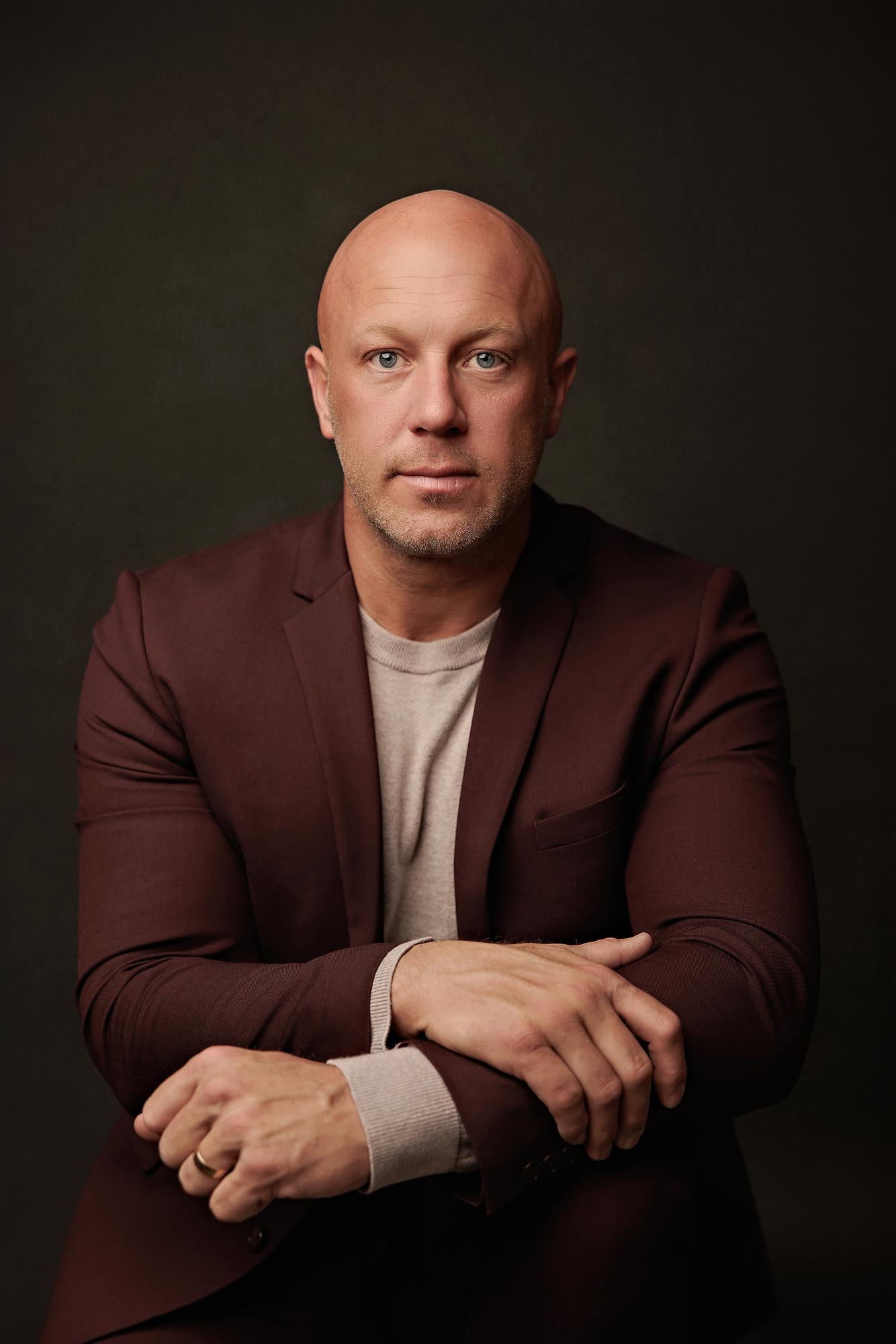 Professional headshot Calgary studio: Close-up portrait of a male executive in a deep burgundy blazer and grey t-shirt leaning forward with arms crossed and a direct, confident gaze against a dark charcoal background